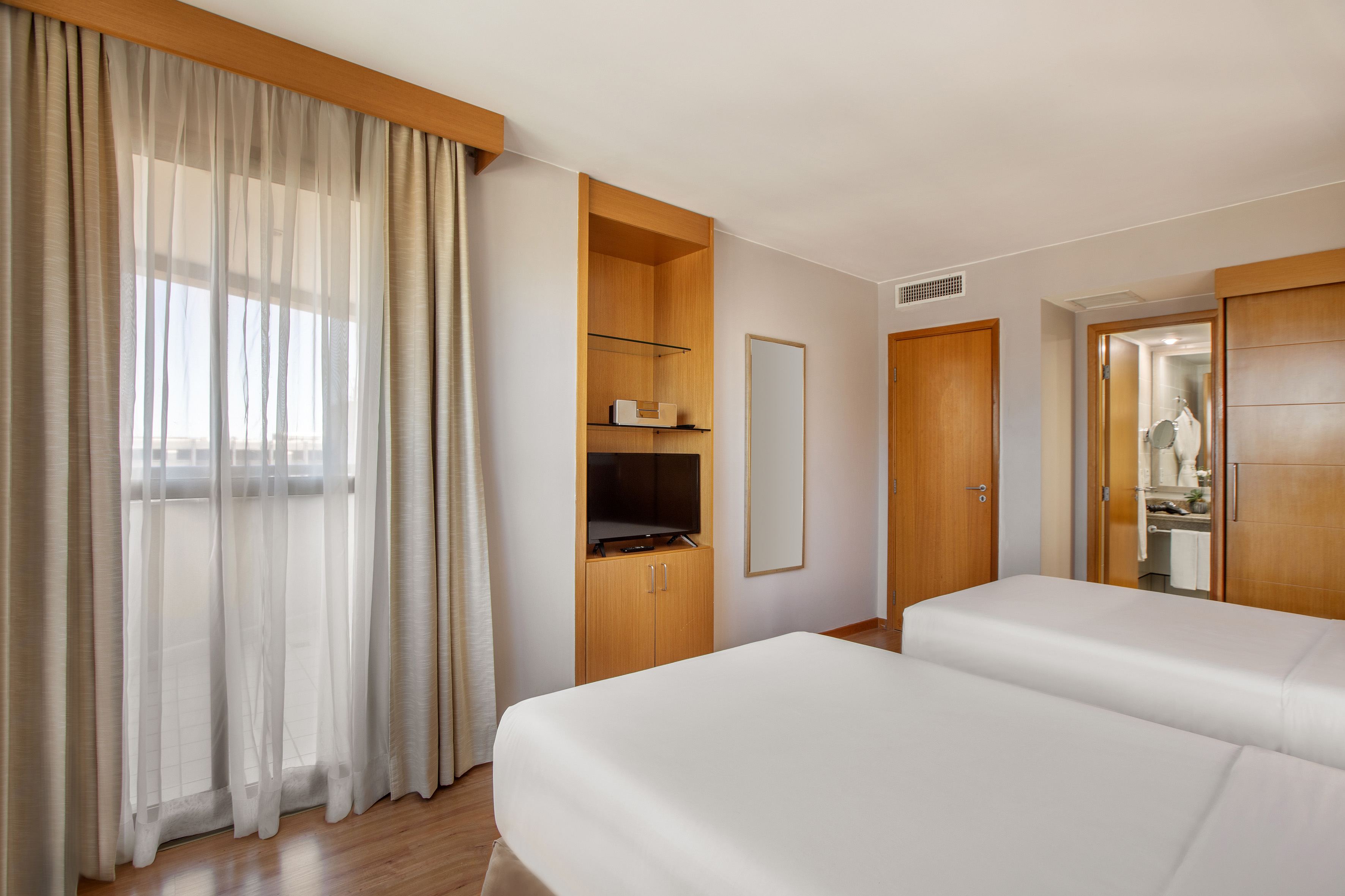 Modern hotel room with two twin beds, TV, and large window with curtains.