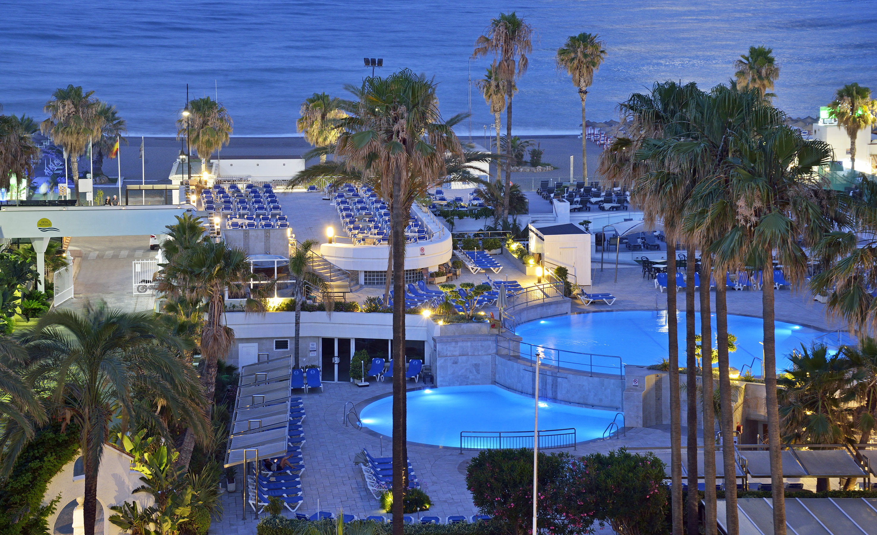 Evening view of a beachfront hotel resort with illuminated pools and palm trees.