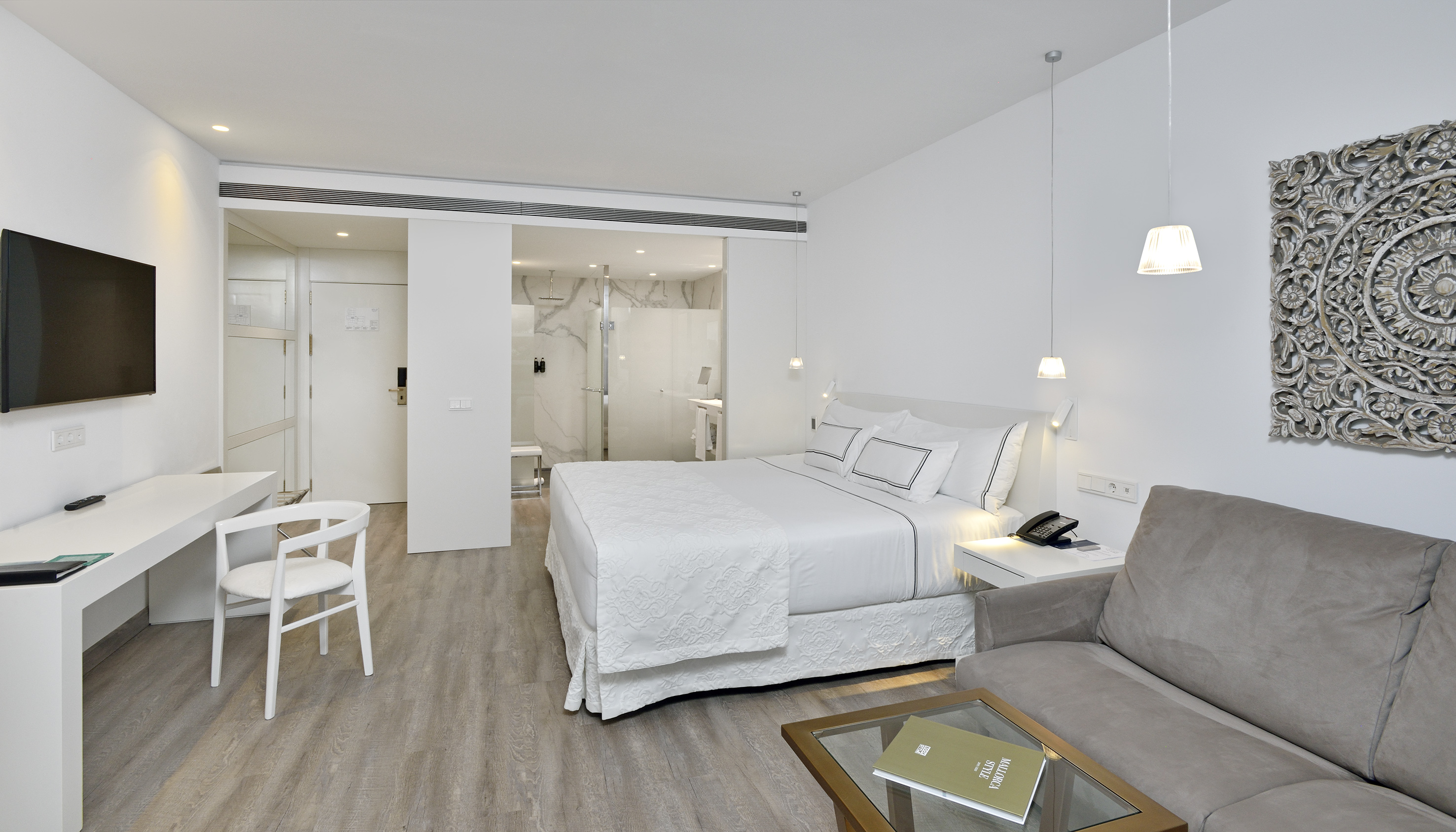Modern hotel room with king bed, white decor, grey sofa, and open marble bathroom.