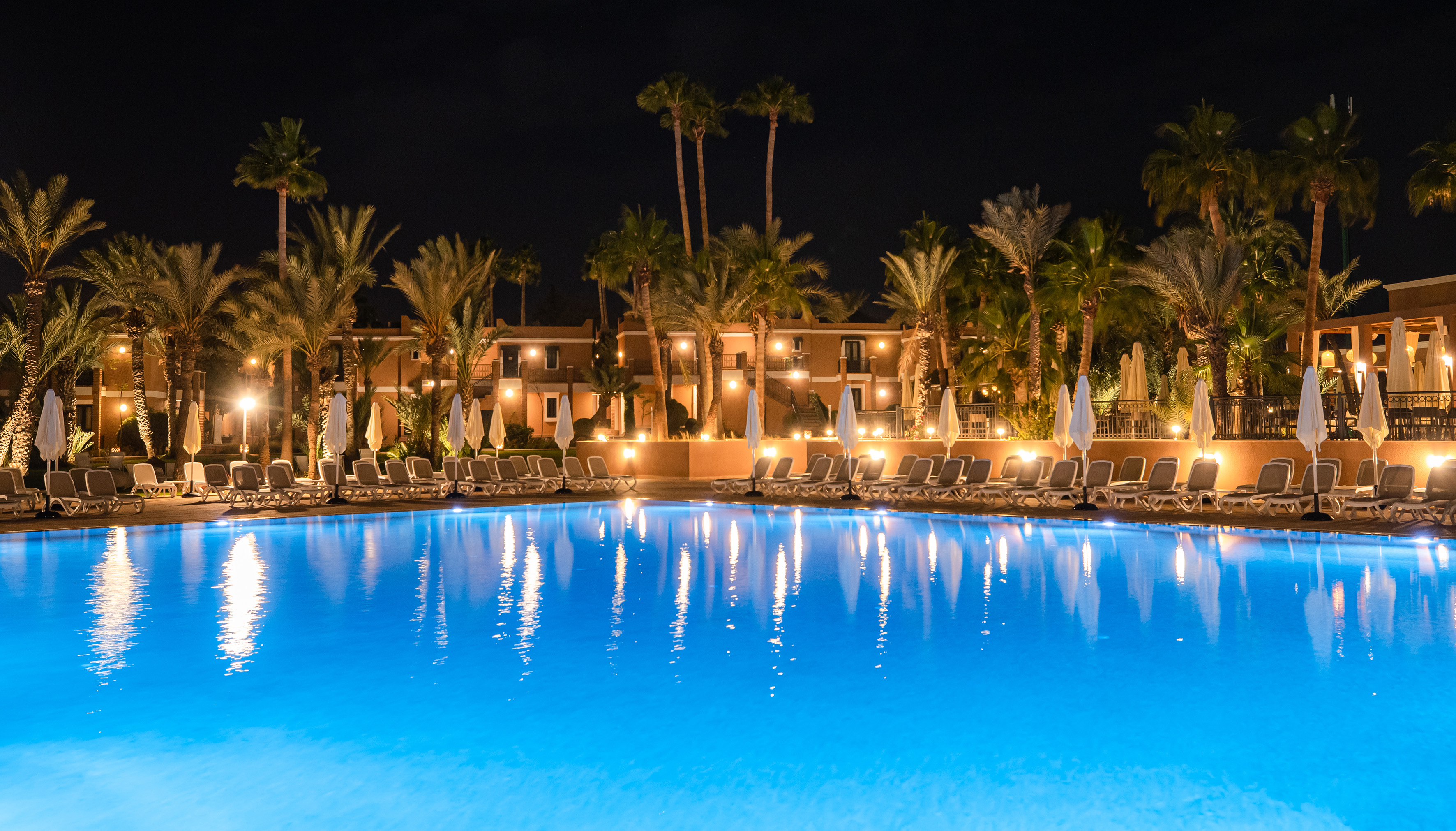 Illuminated resort pool at night with palm trees, loungers, and bright buildings.