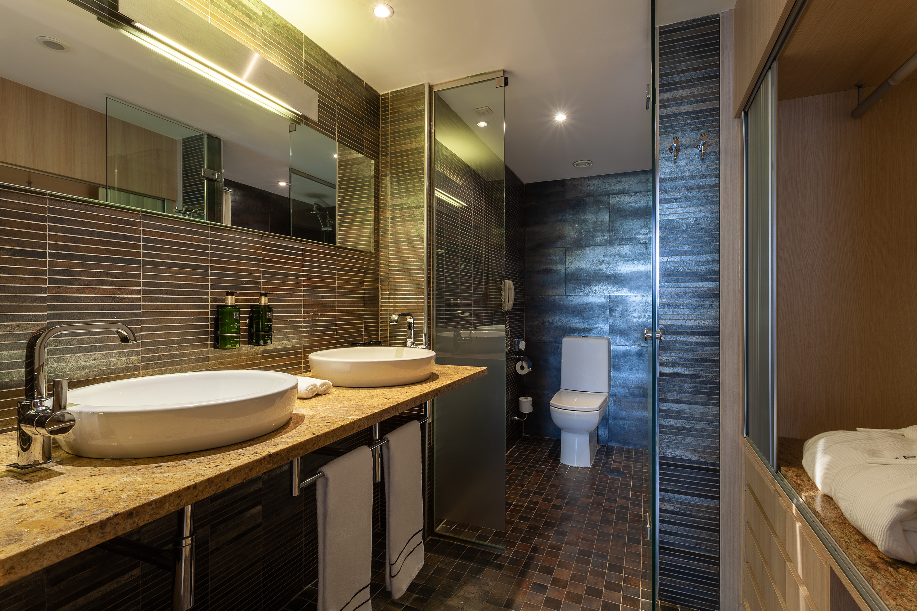 Hotel bathroom with dual sinks, granite vanity, dark tiles, and toilet.