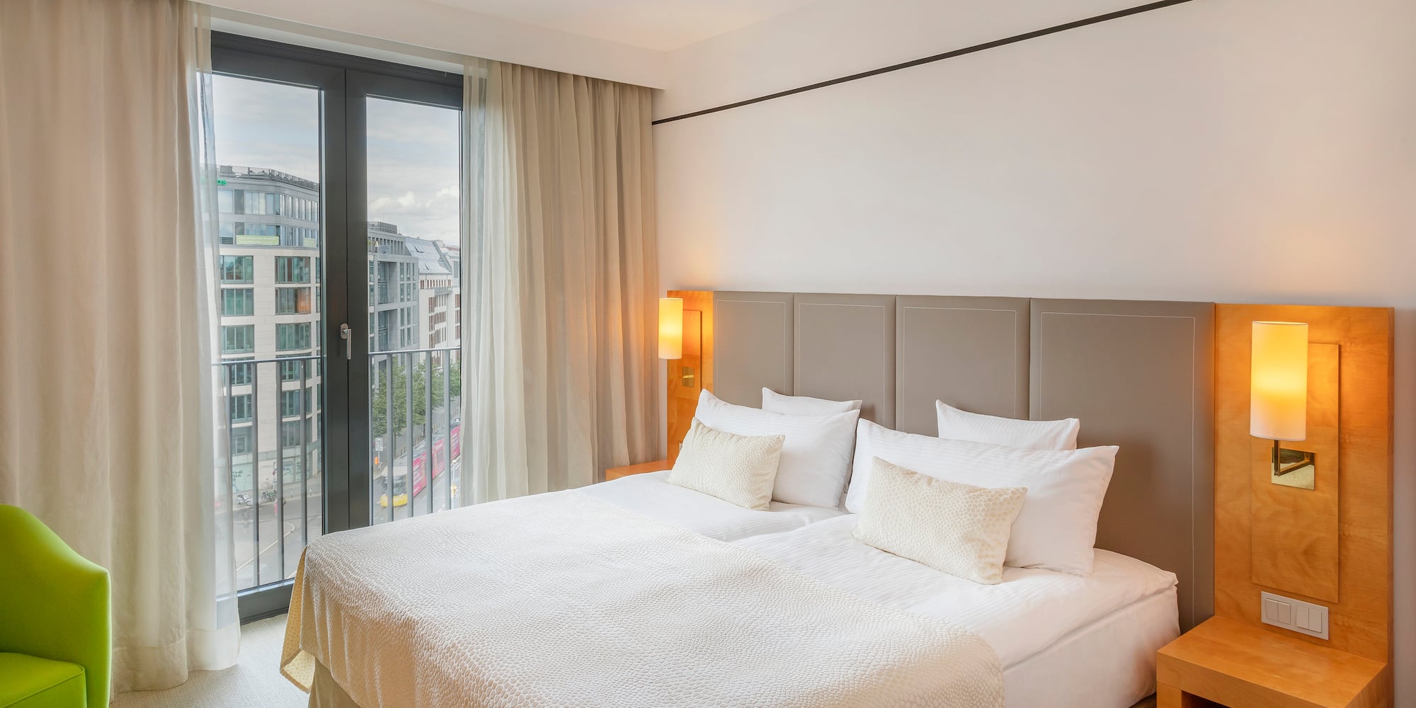 Modern hotel bedroom with large bed, cityscape from window, and green armchair.