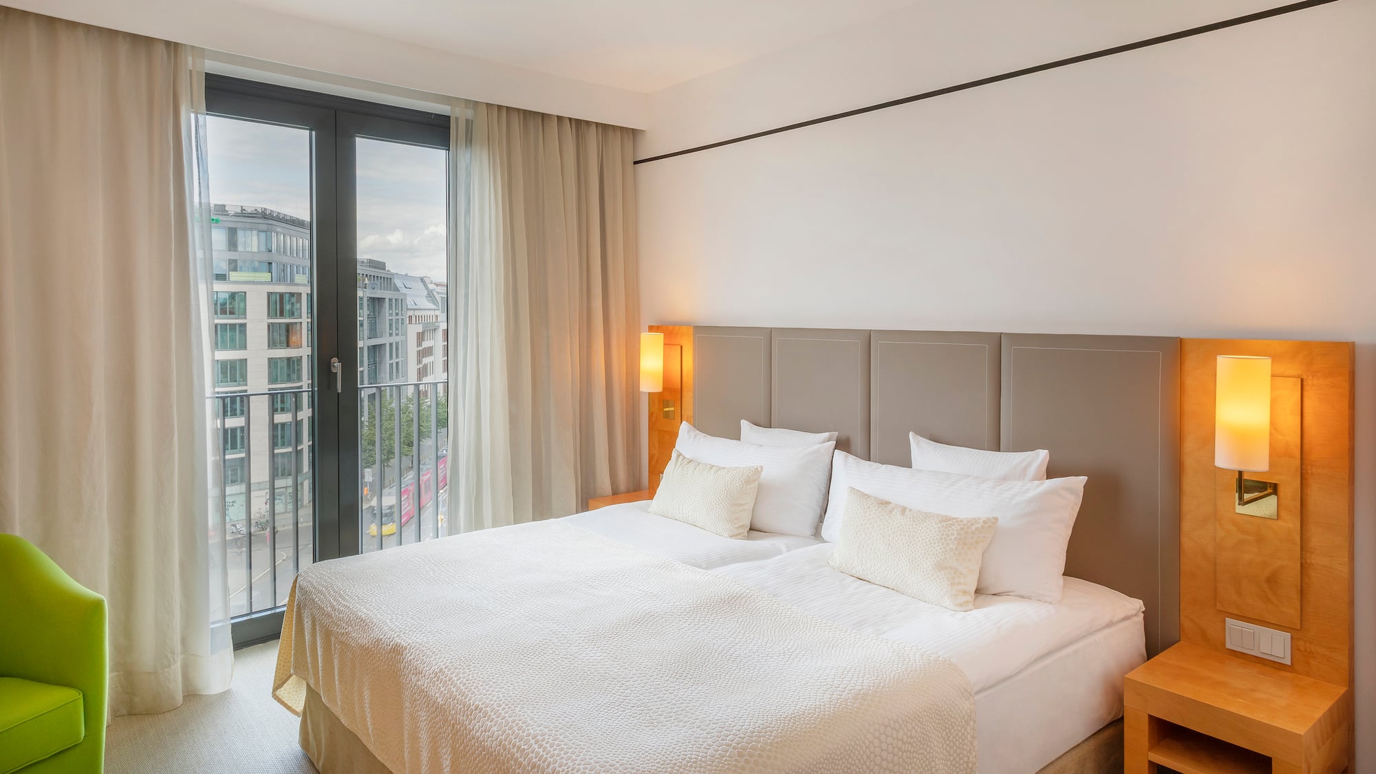 Modern hotel bedroom with city view, large bed, and green armchair.