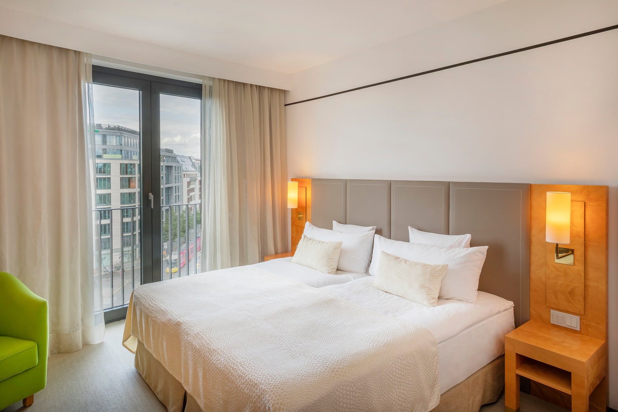 Modern hotel room with king bed, window city view, and green accent chair.