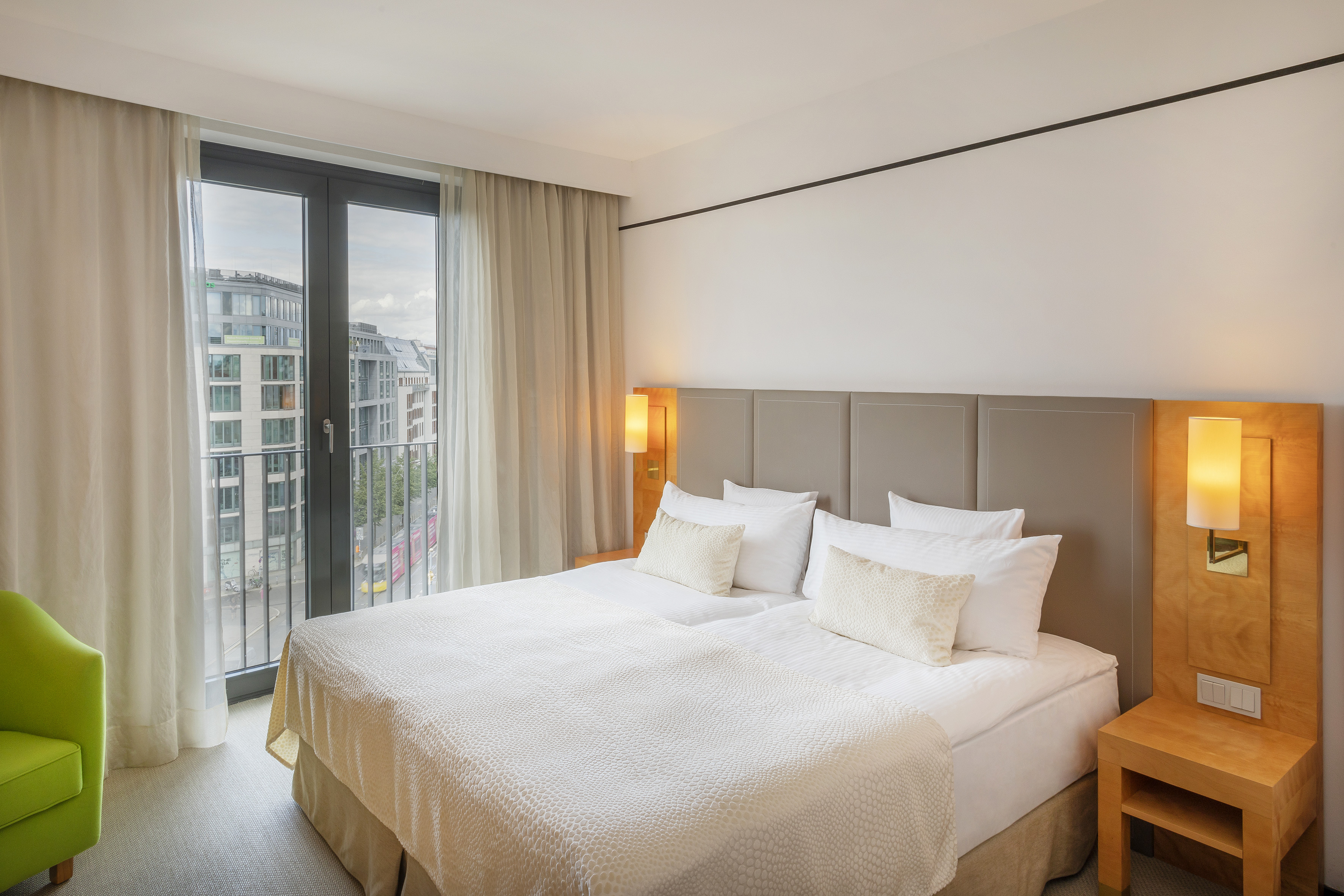 Modern hotel bedroom with large bed, cityscape from window, and green armchair.