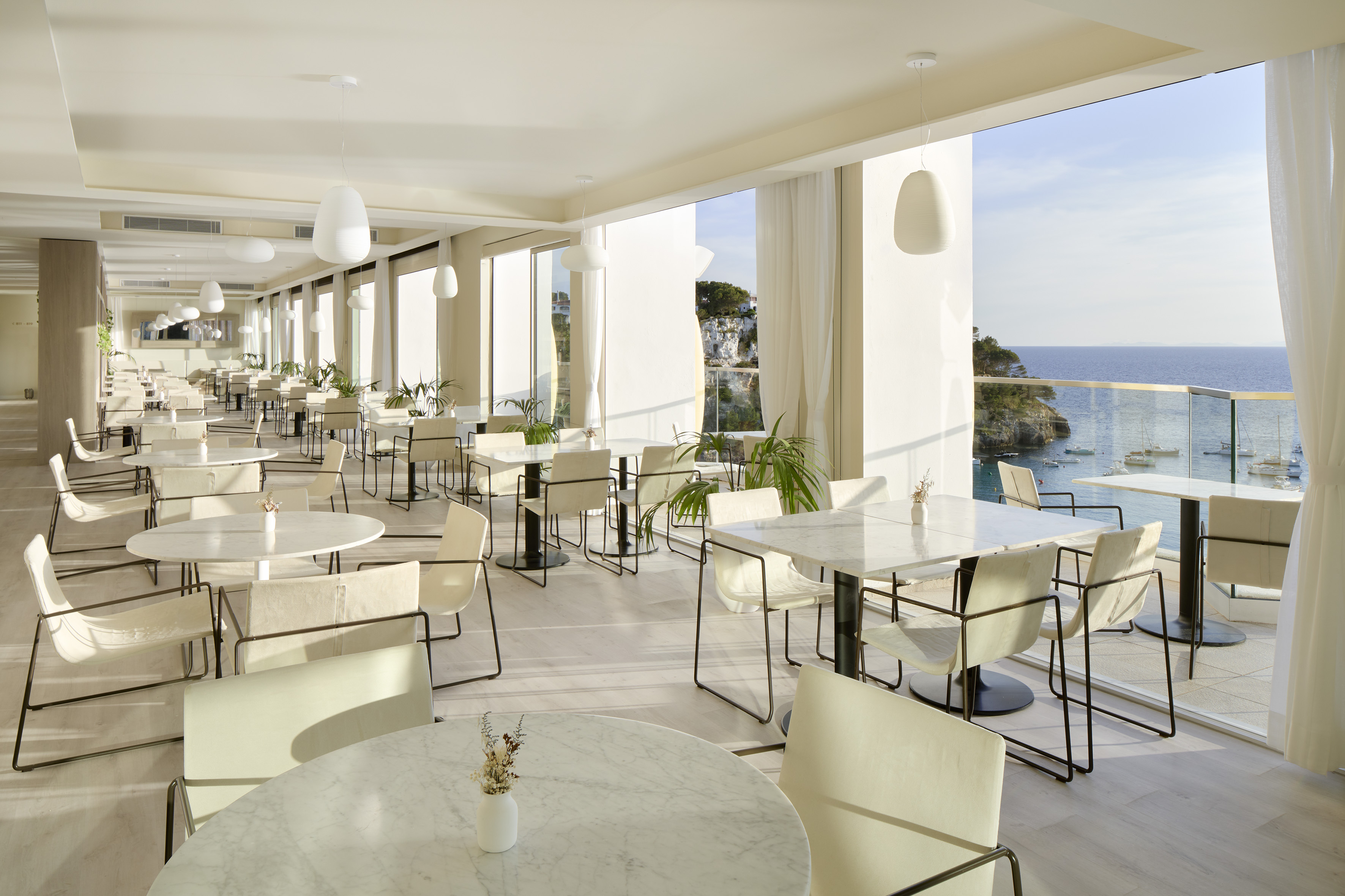 Modern restaurant with panoramic sea view, white tables, and coastal scenery.