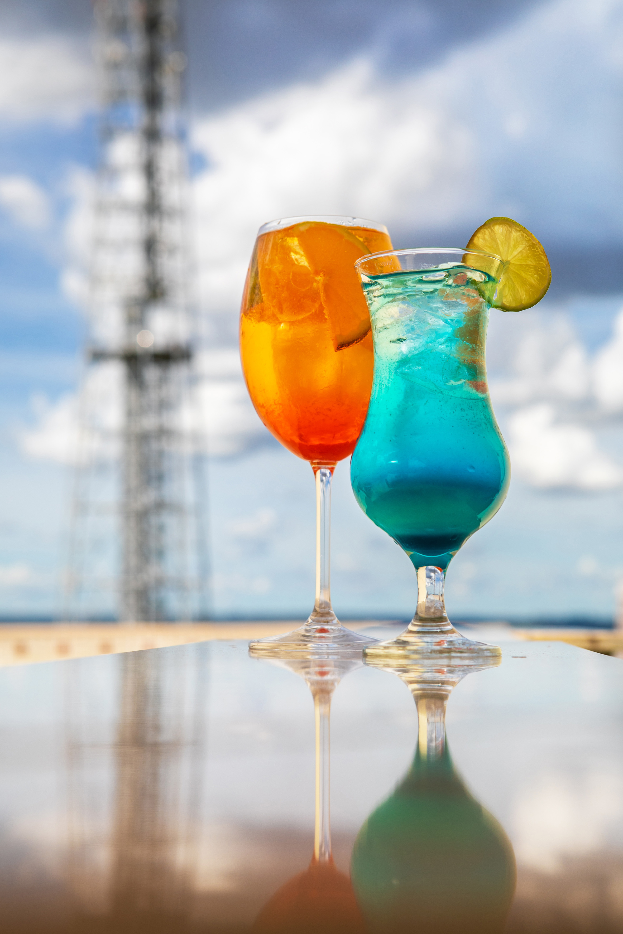 Orange Aperol Spritz and blue cocktail with lime on reflective outdoor table.