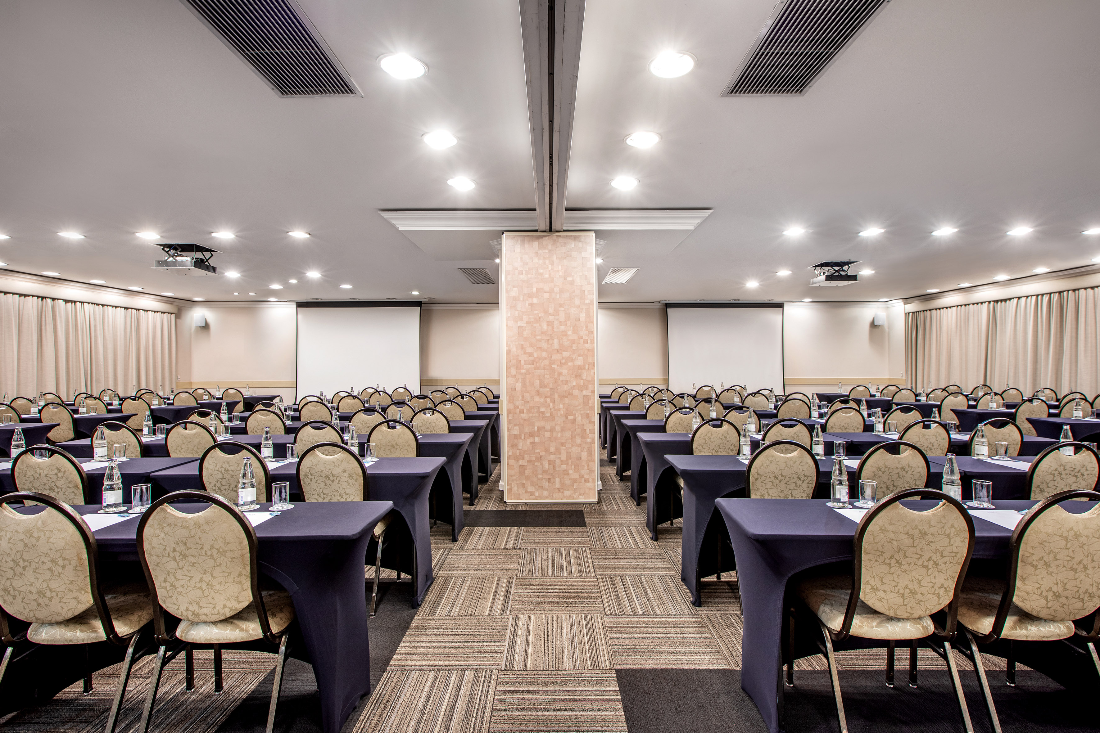 Conference room with rows of tables, chairs, water, and dual projection screens.