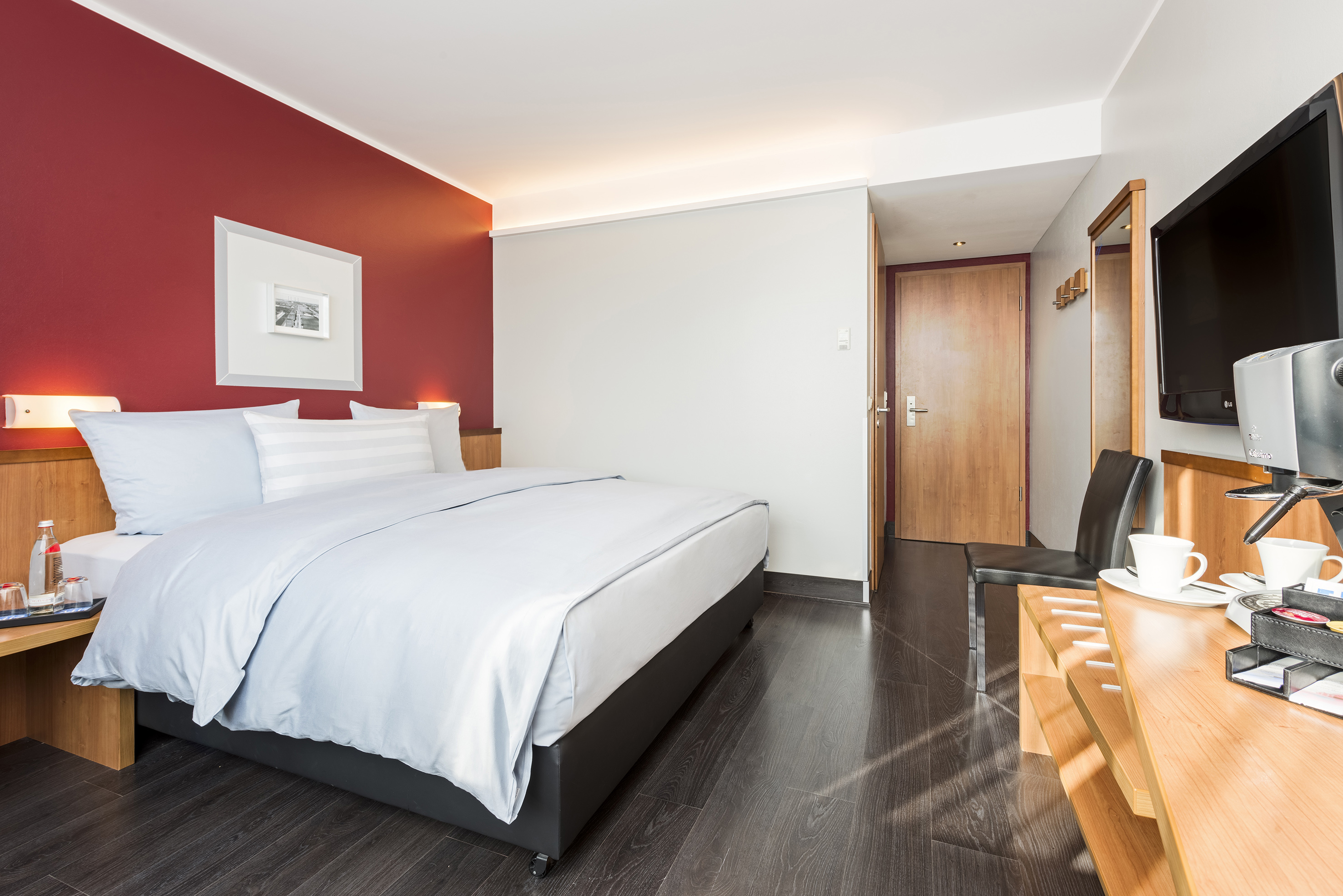 Modern hotel room with a bed, red accent wall, dark wood floor, TV, and coffee maker.