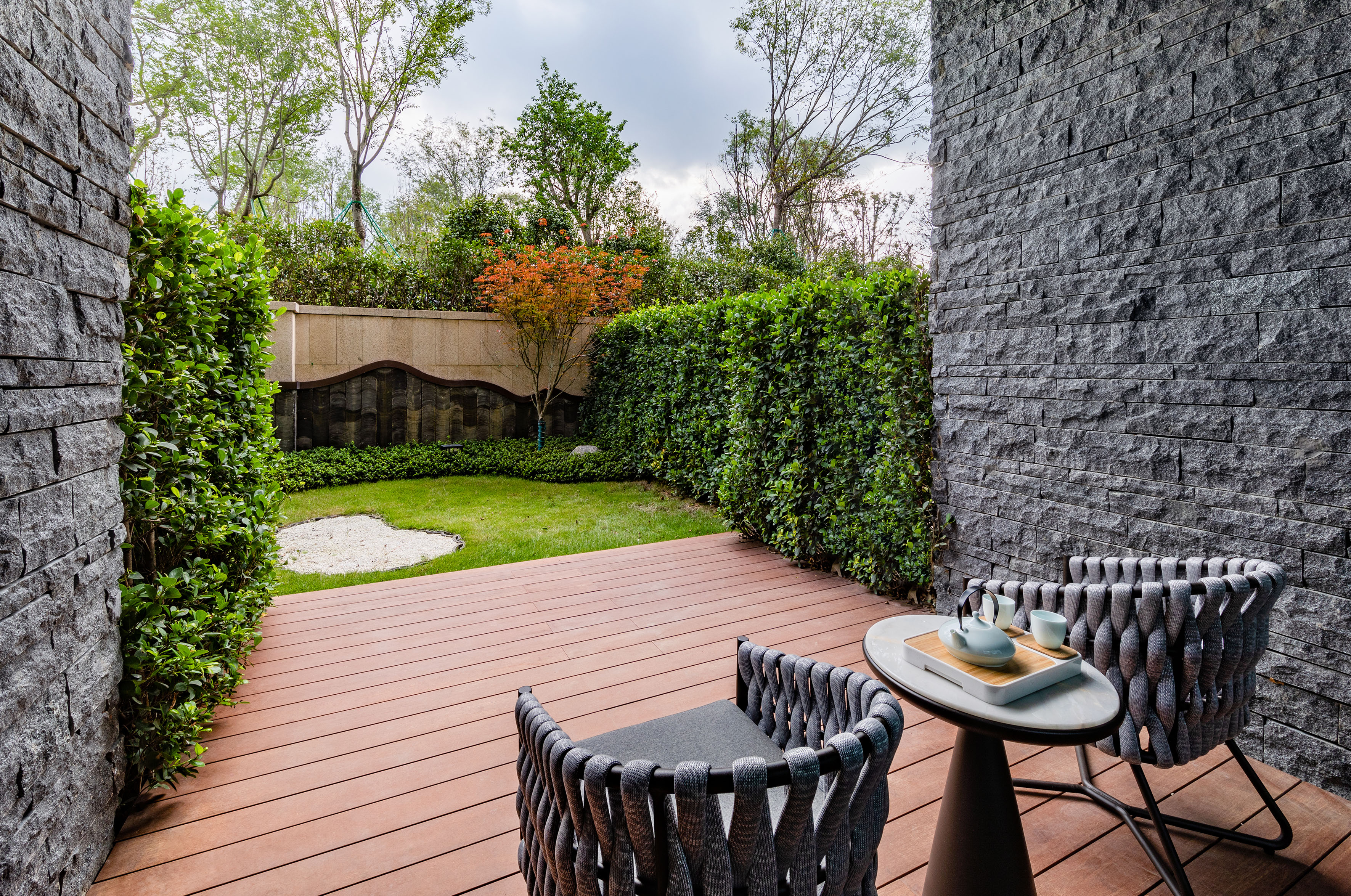 Modern patio with wood deck, chairs, tea set, stone wall, and green garden.