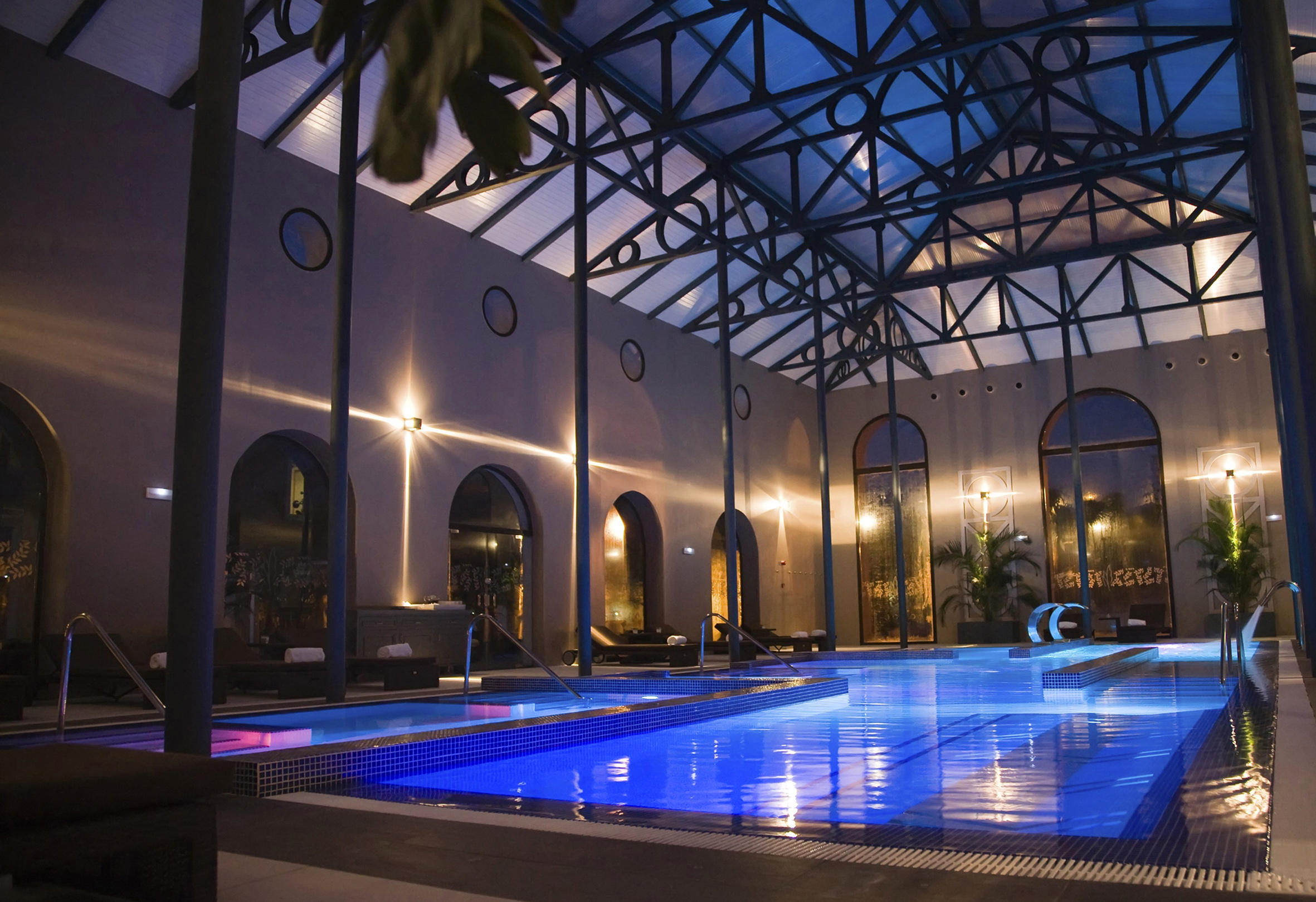 Illuminated indoor spa pool with blue water, glass roof, and arched windows.