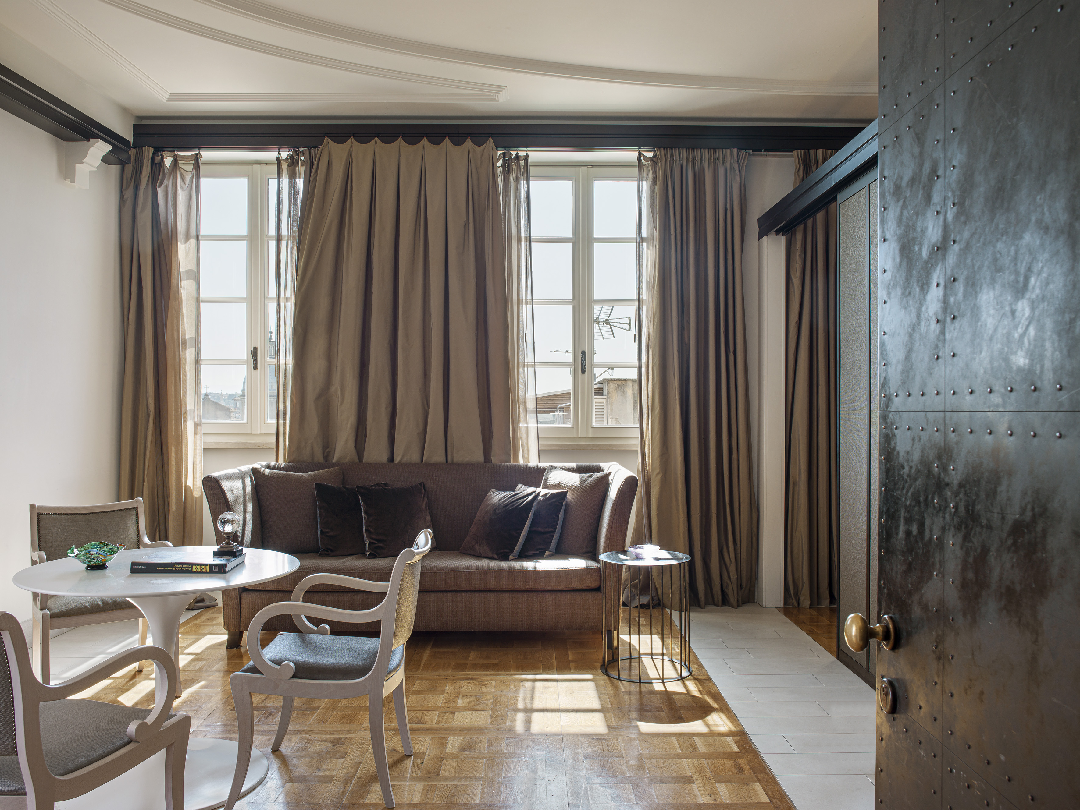 Living room with brown sofa, white table, parquet floor, bright windows, and studded door.