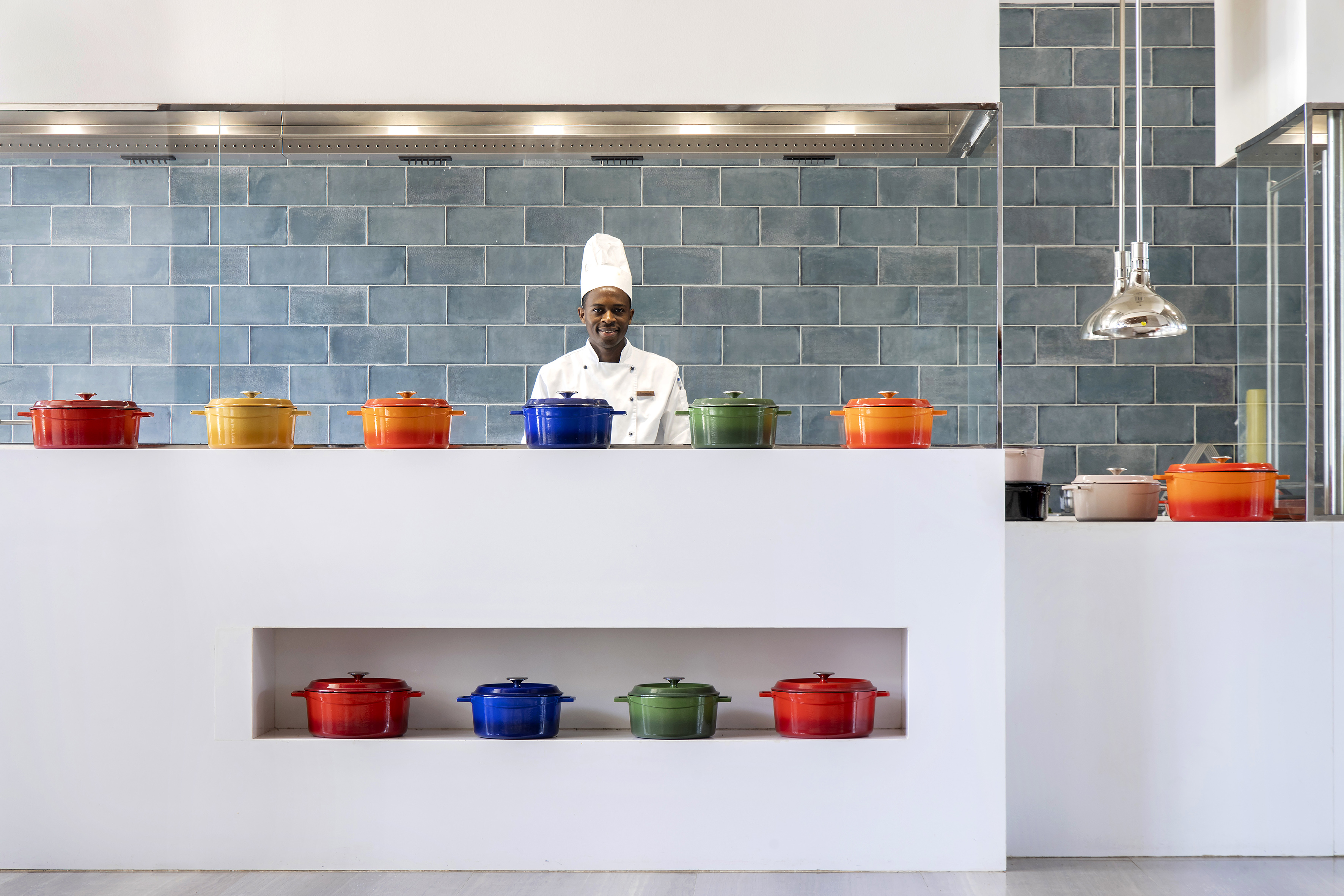 Smiling chef with colorful cooking pots in a modern, blue-tiled kitchen.