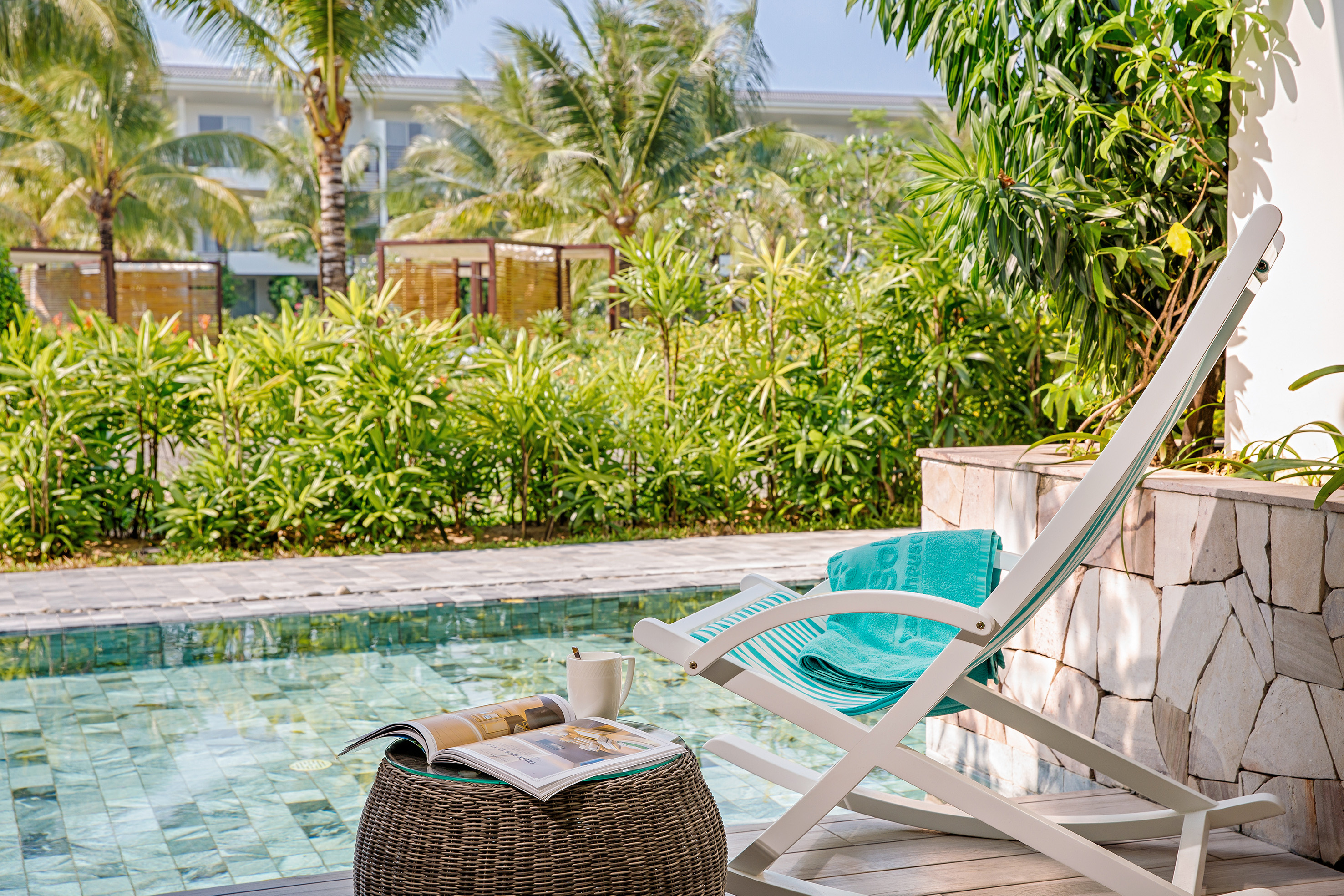 Tropical resort pool deck with lounge chair, magazine, coffee, and palm trees.