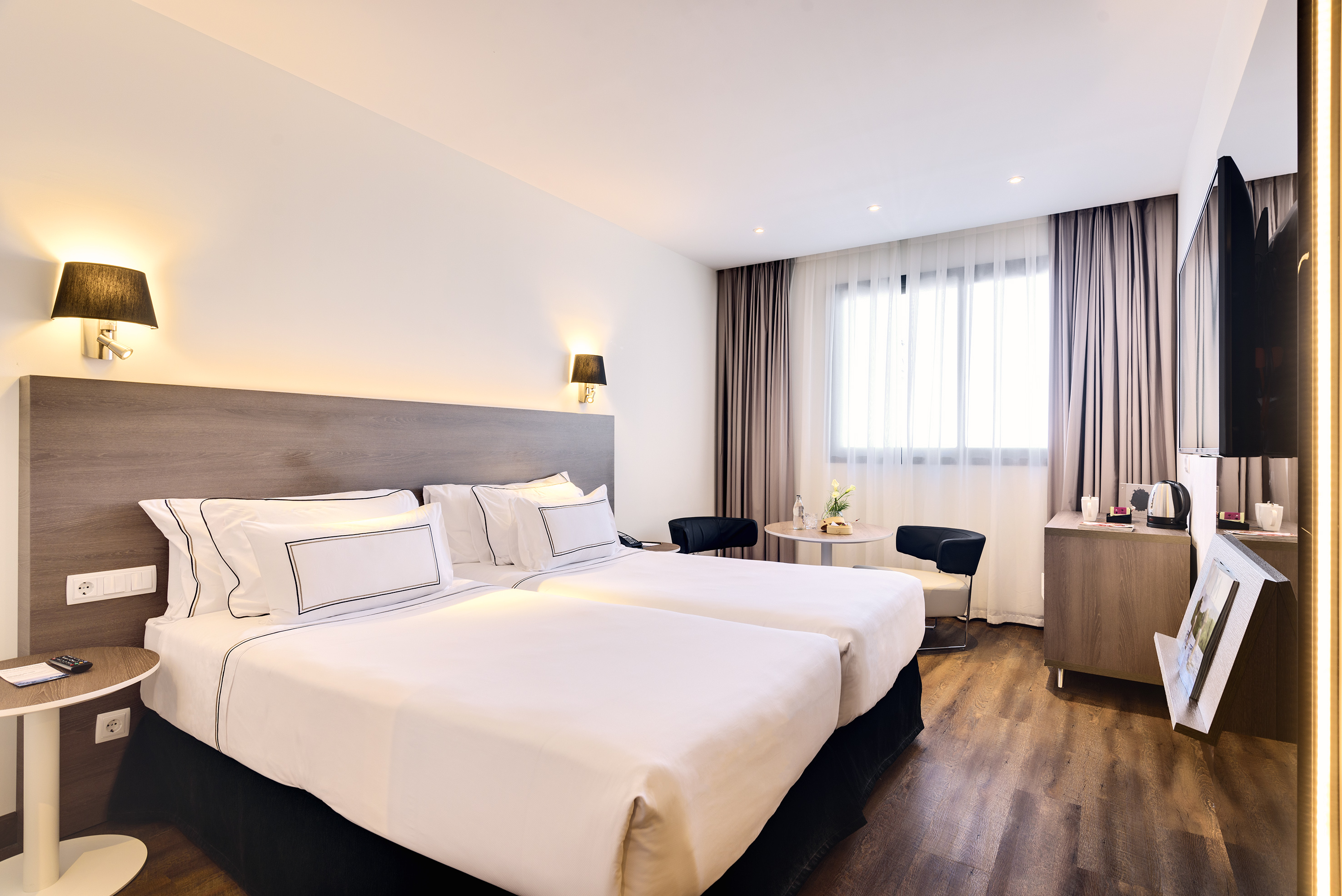Modern hotel room with two twin beds, wooden floor, and bright window.