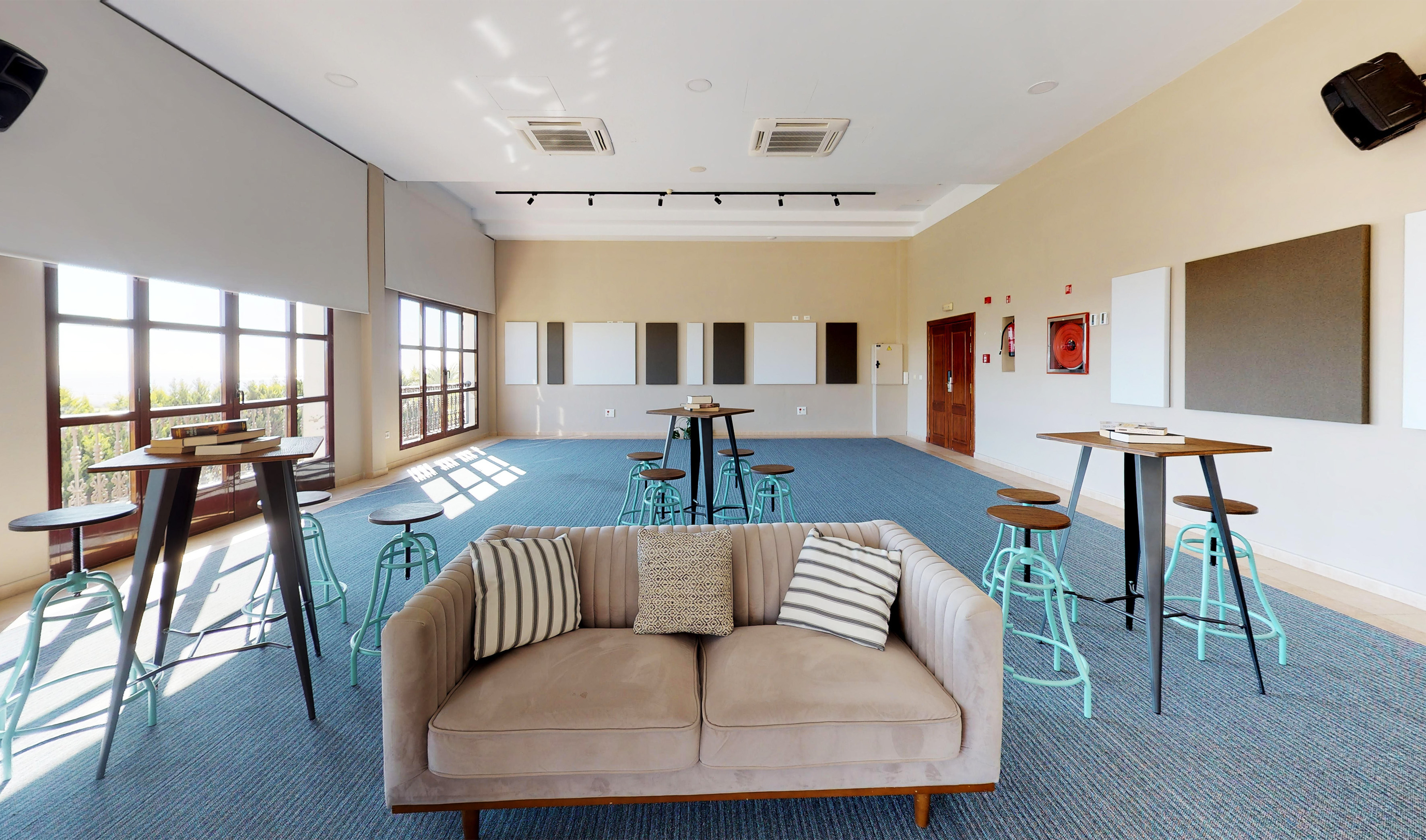 Modern meeting lounge with blue carpet, high tables, sofa, and sunlit windows.