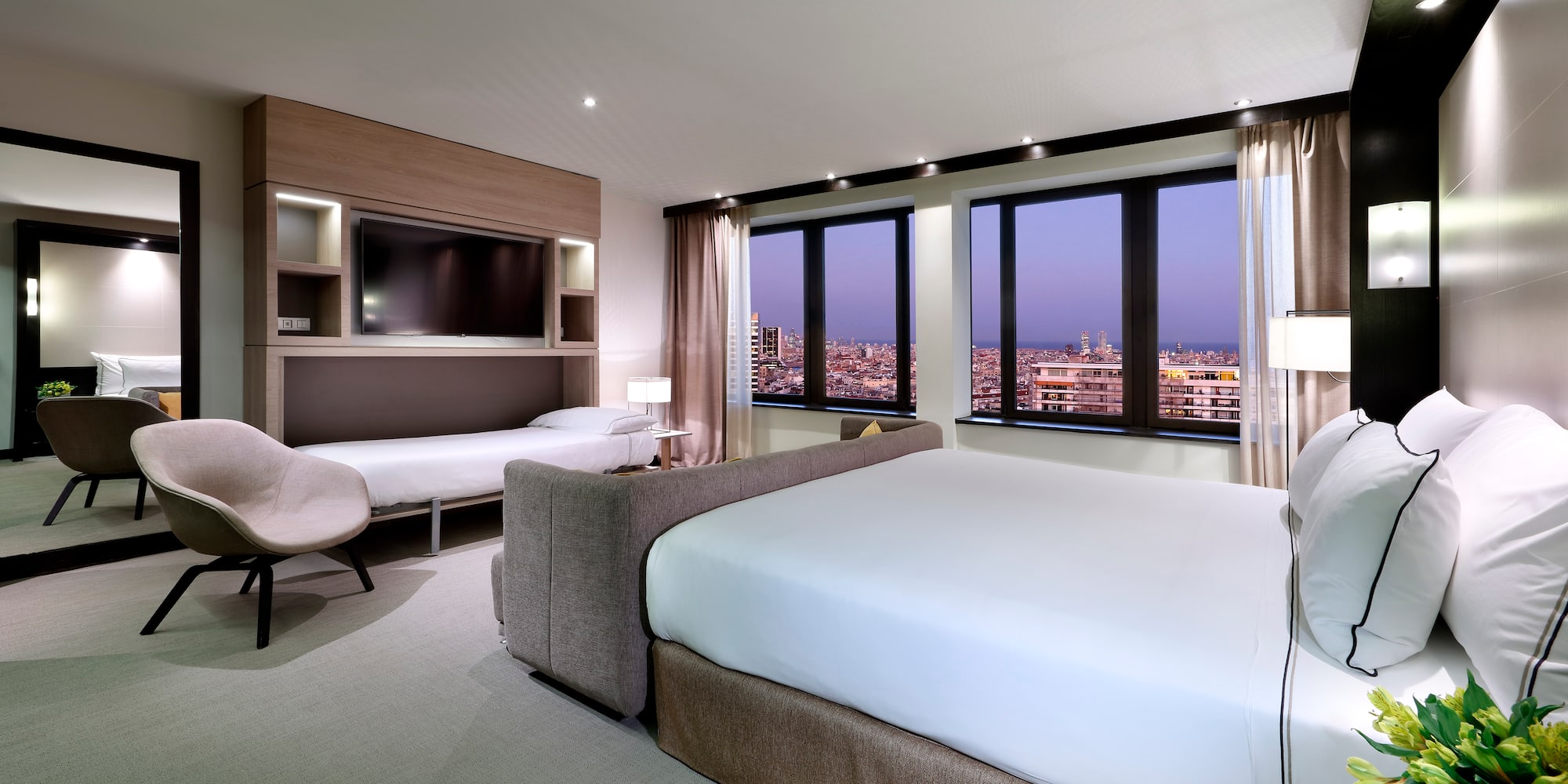 Modern hotel room with city night view, large bed, and integrated fold-down bed.