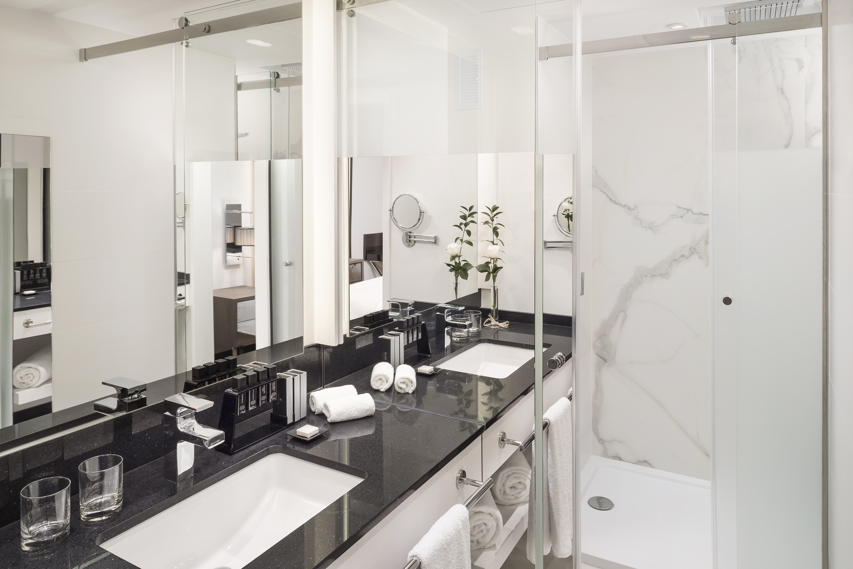 Modern hotel bathroom with double vanity, black granite counter, and walk-in shower.