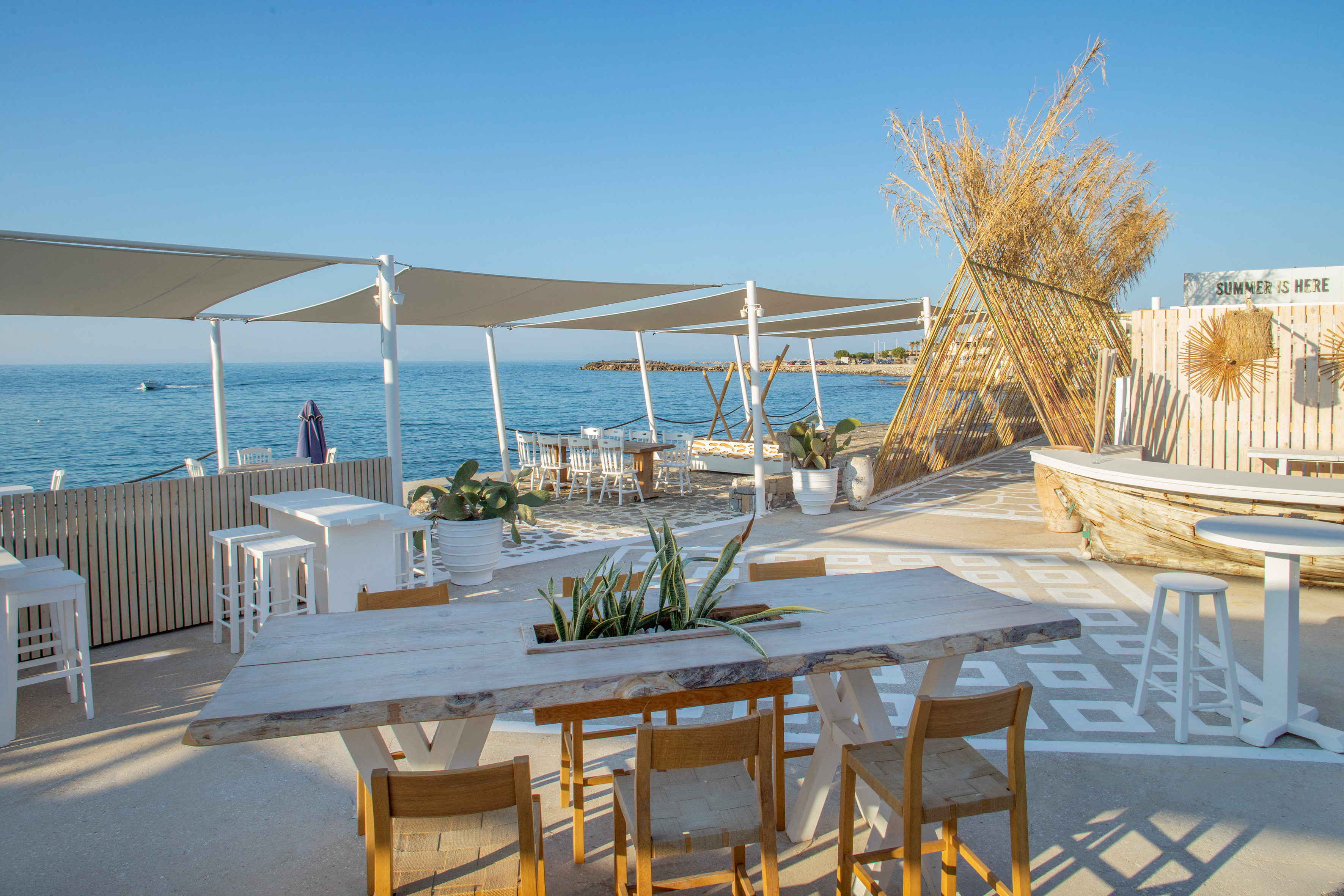 Beachfront restaurant with white tables, shade sails, and clear blue sea.