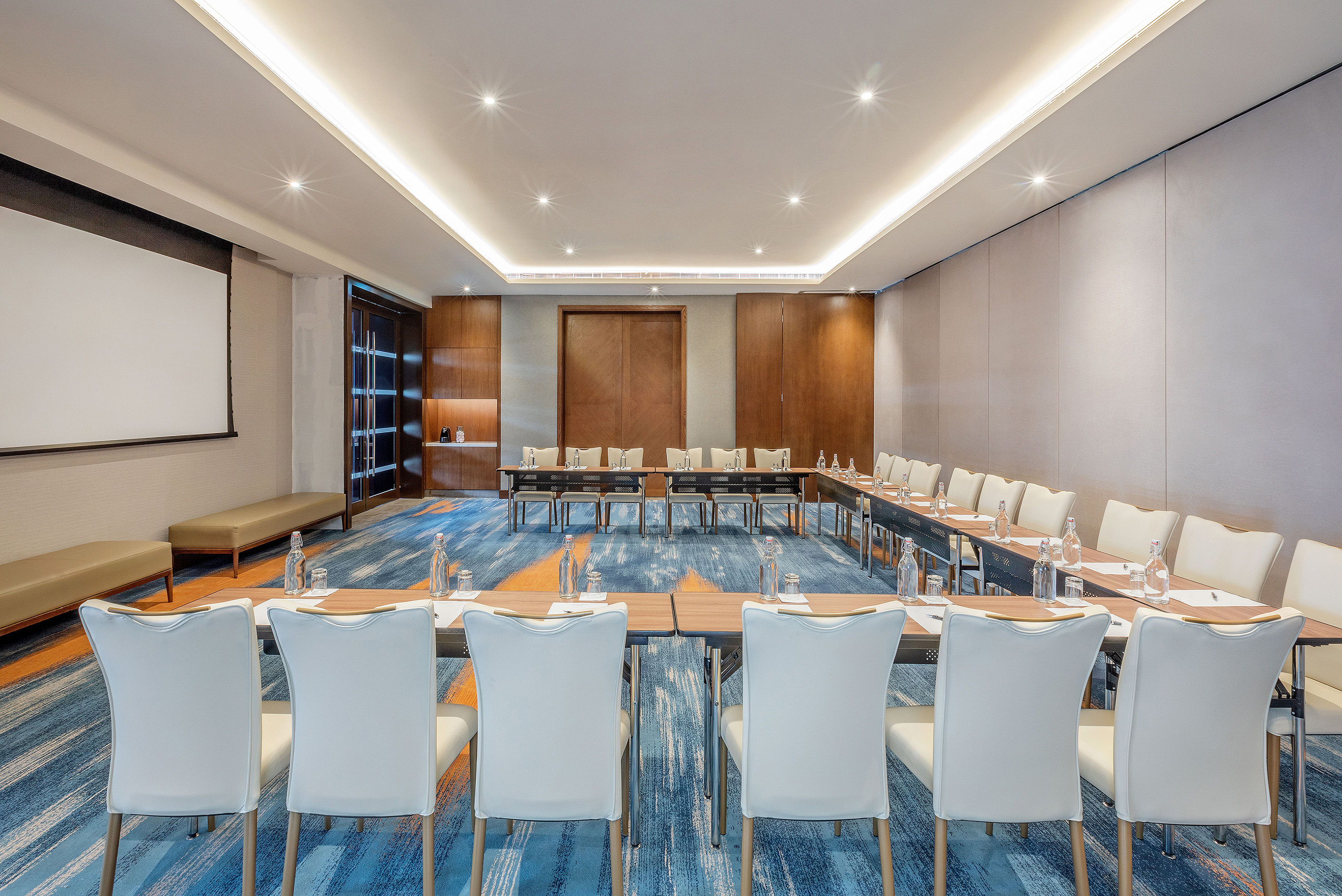 Modern U-shaped conference room with white chairs, projector screen, blue carpet.