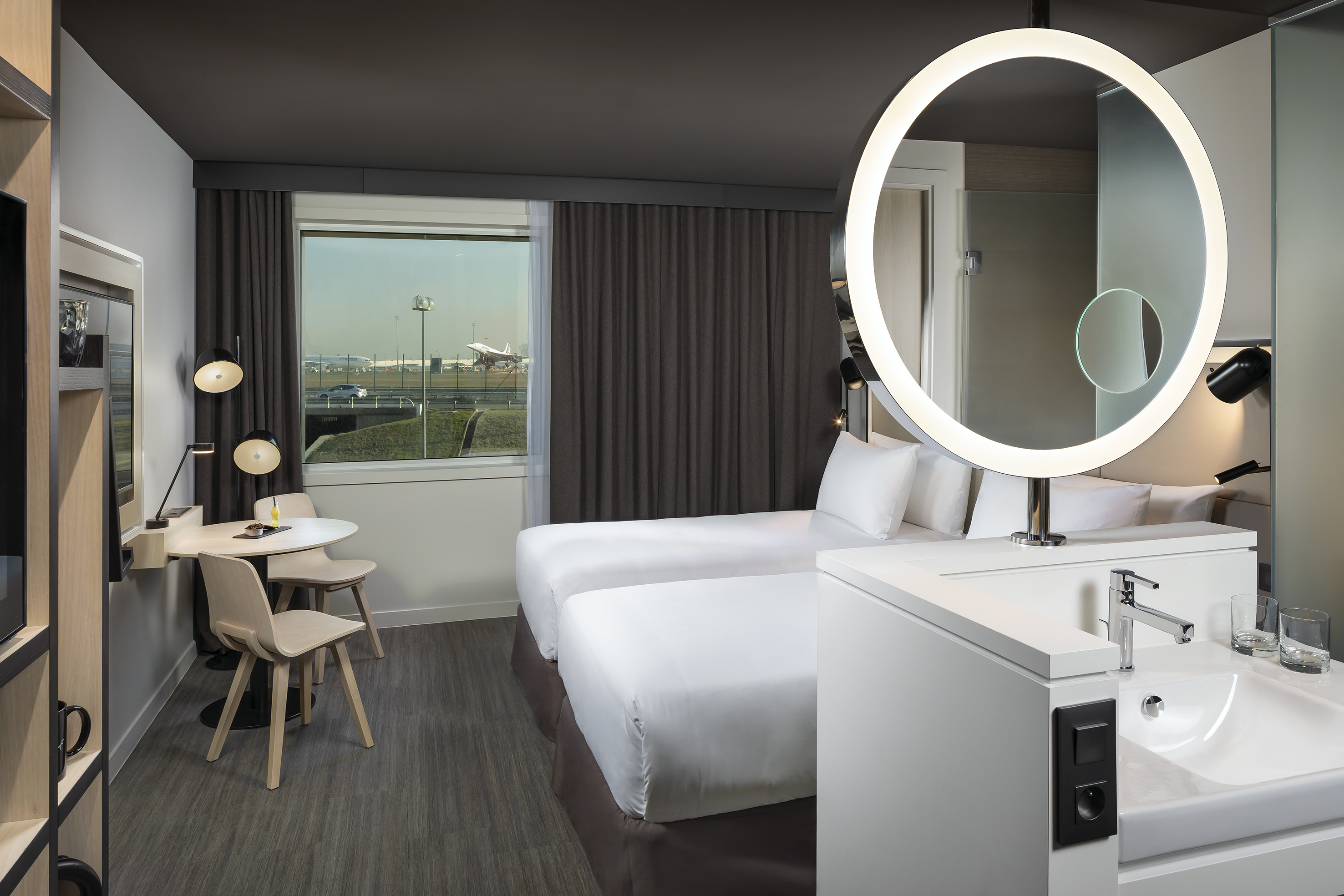 Airport hotel room with twin beds, illuminated mirror, and runway view.