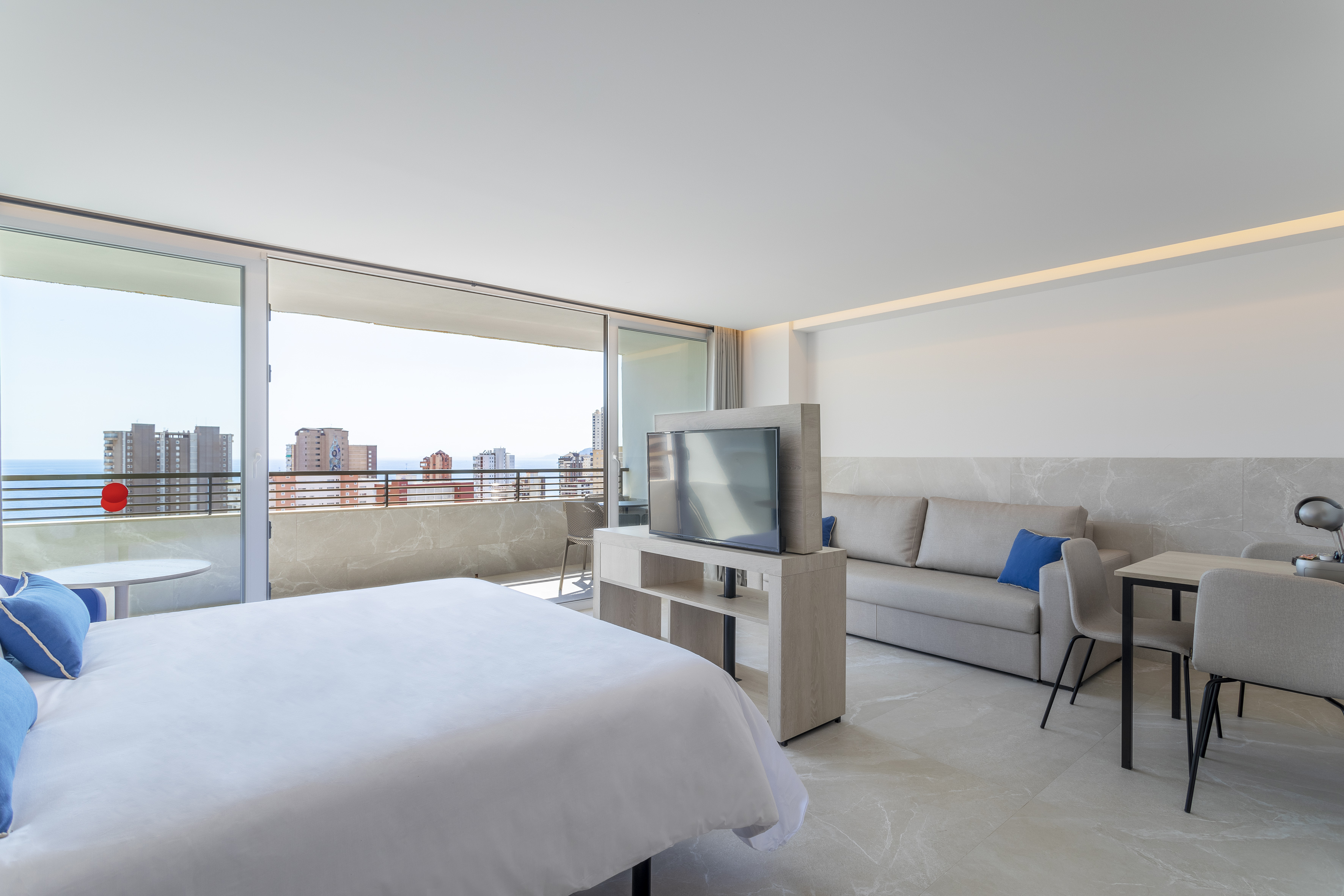 Modern hotel room with ocean view balcony, bed, sofa, and city skyline.