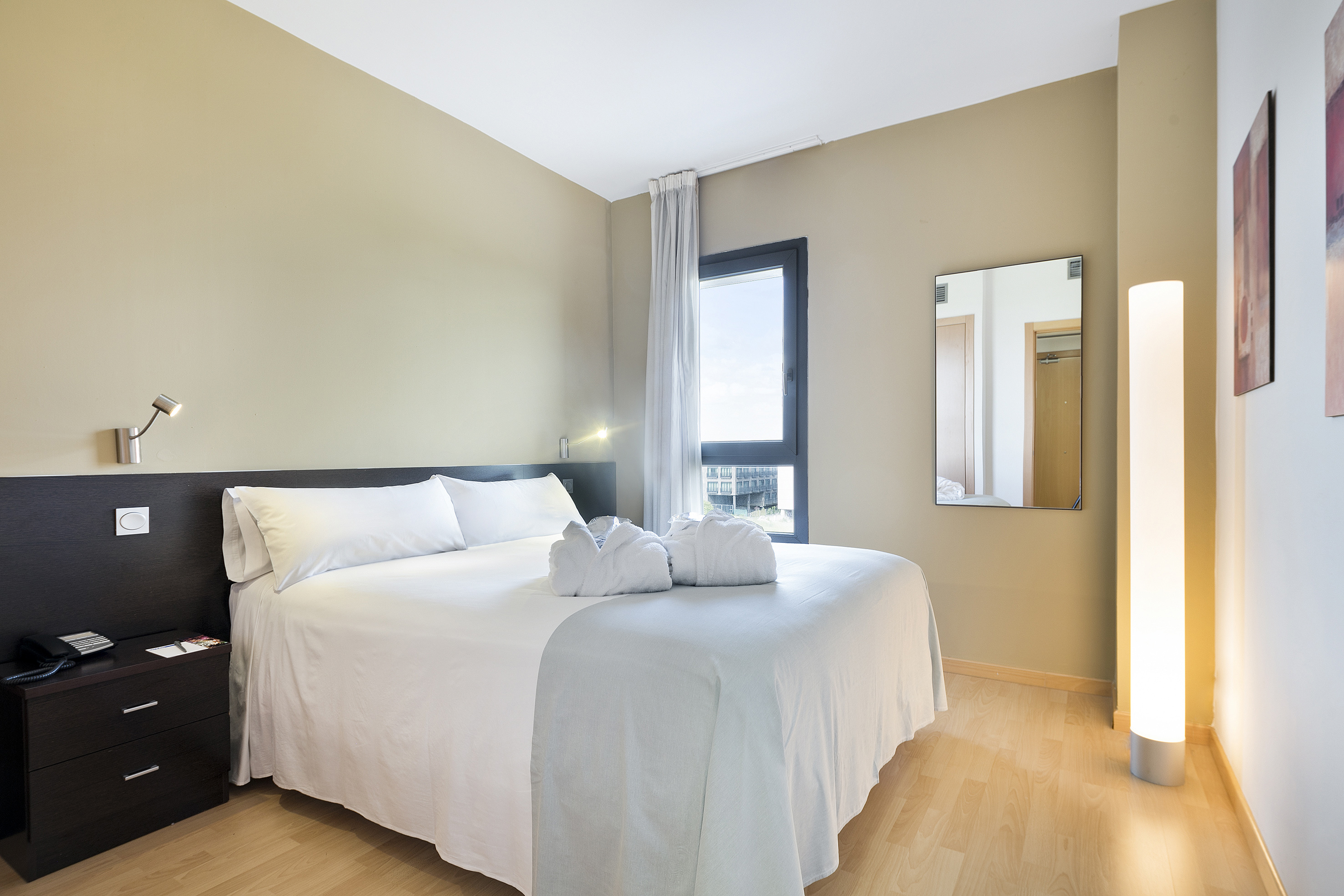 Modern hotel bedroom with white bed, bathrobes, window, and wood floor.