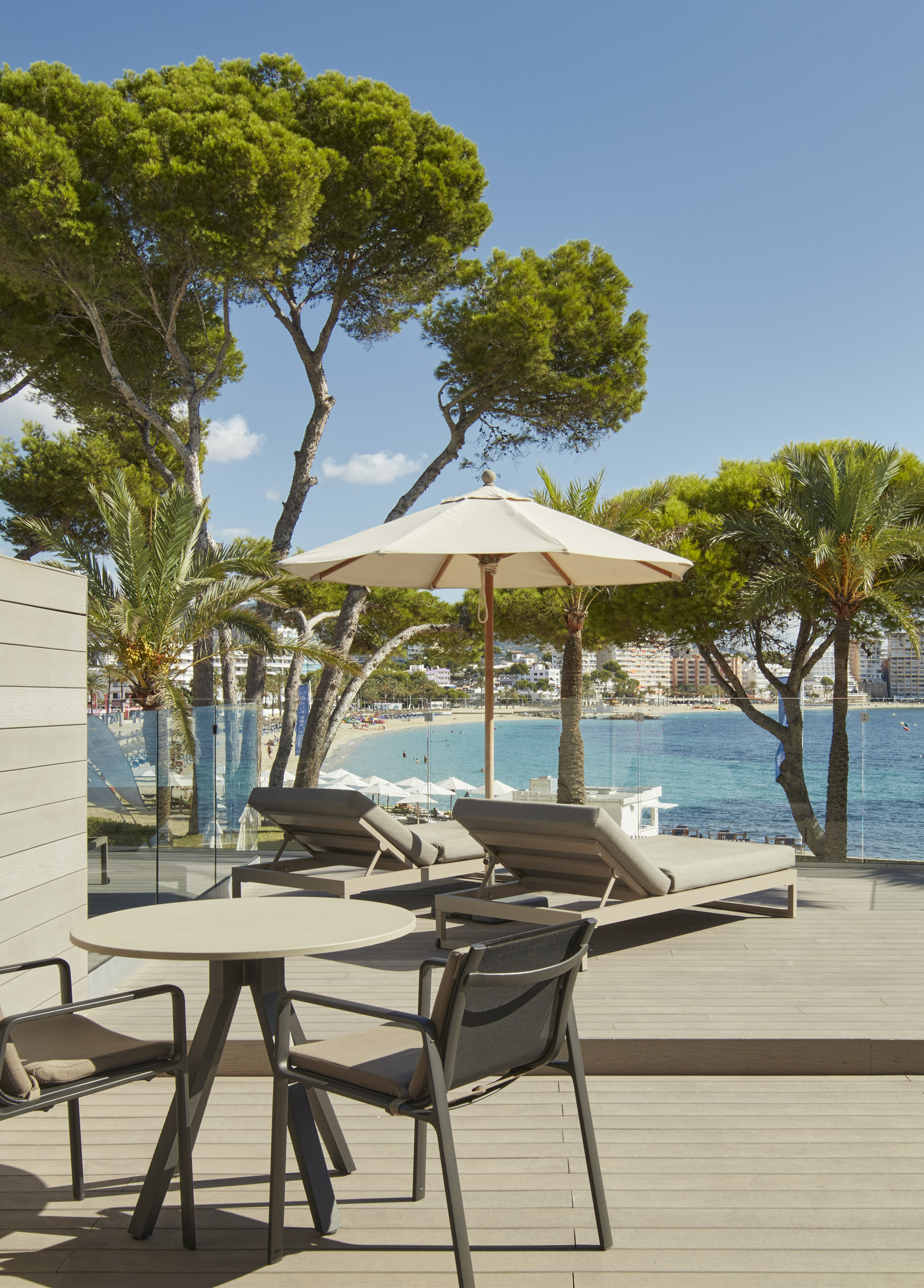 Sunny hotel terrace with sun loungers, pine trees, and a clear blue ocean view.