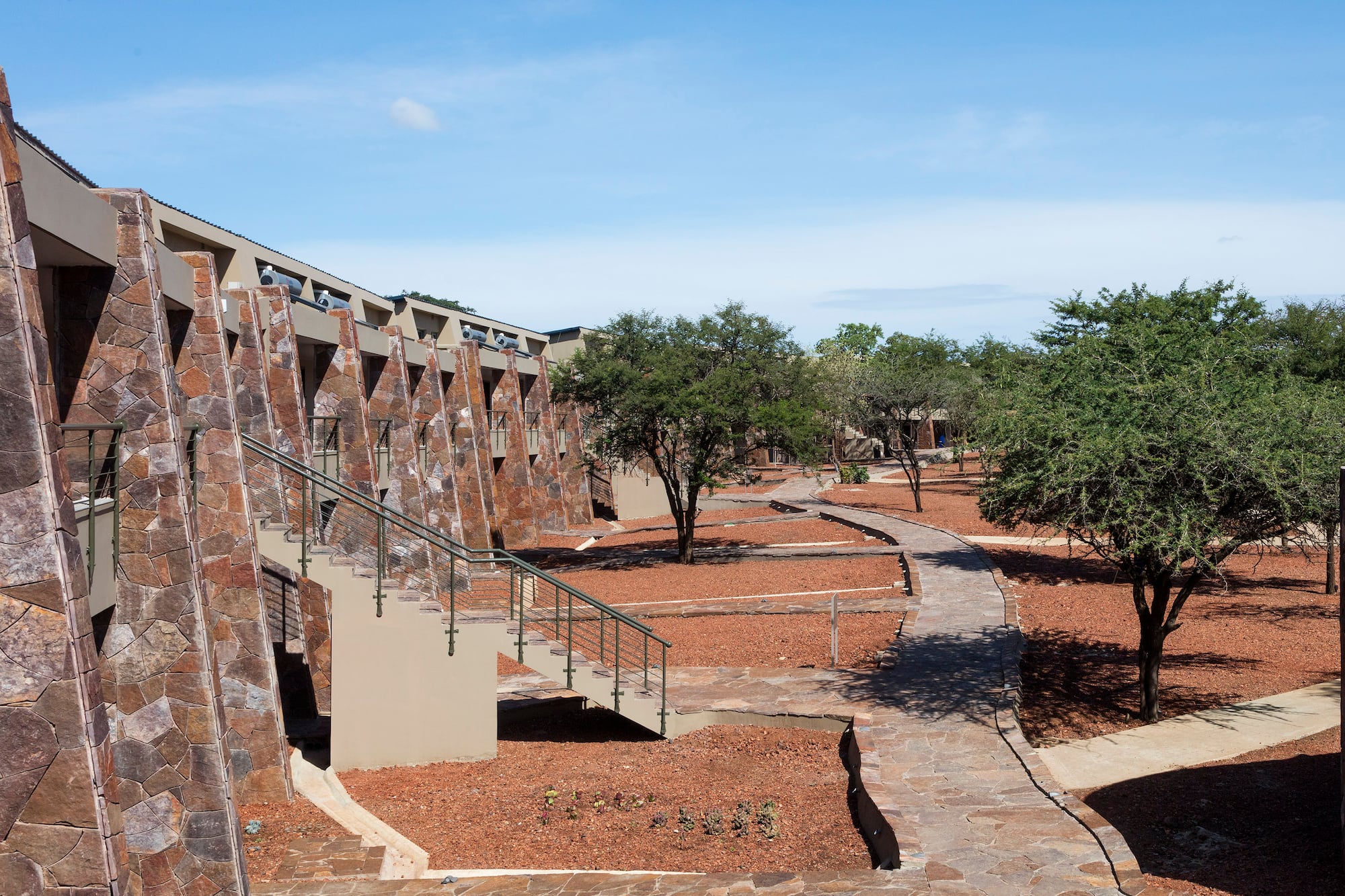 Stone-faced safari lodge, steps, winding paths, red earth, and green trees.