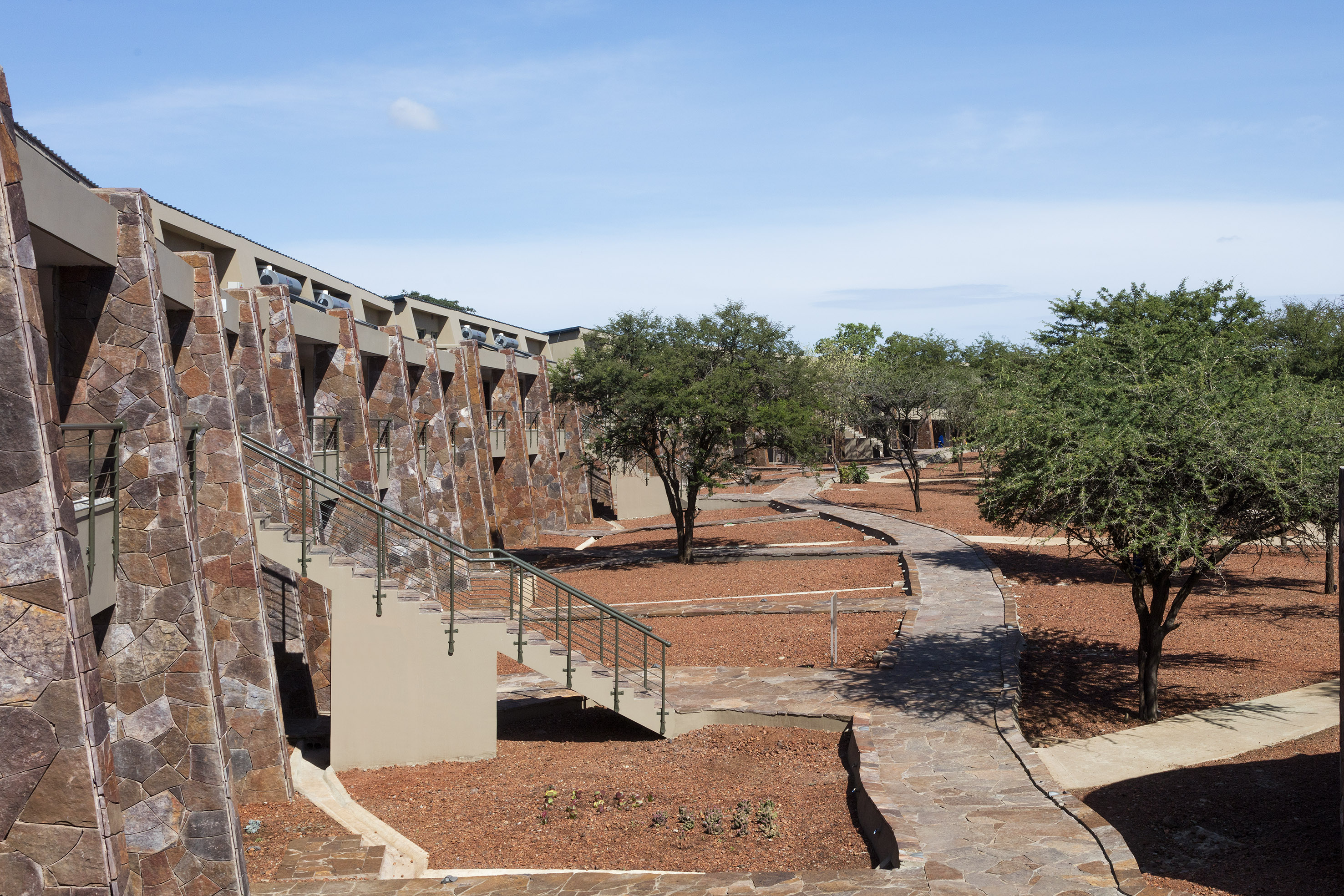 Stone-faced safari lodge, steps, winding paths, red earth, and green trees.
