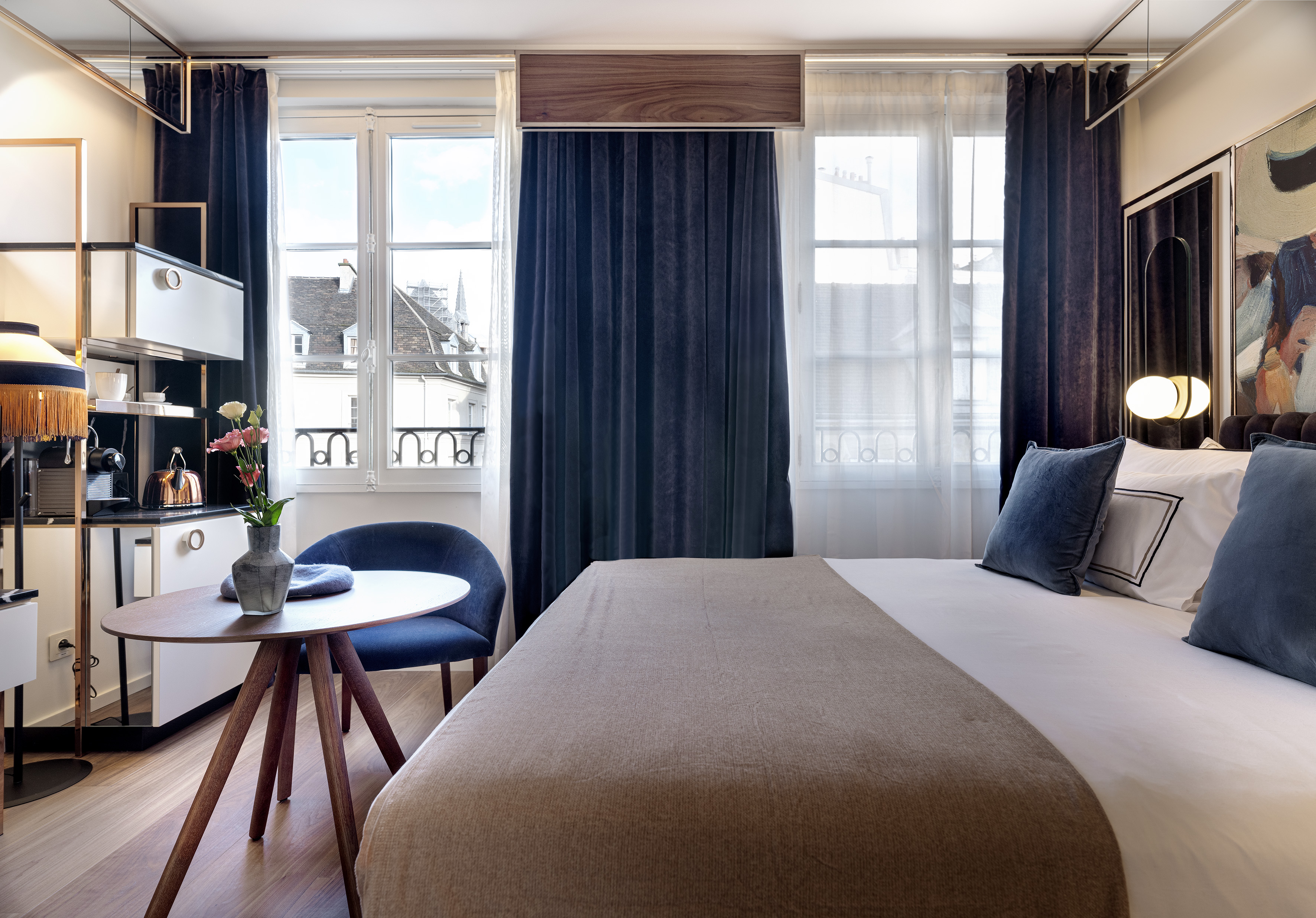 Modern hotel room with bed, city view windows, blue armchair, and mini bar.