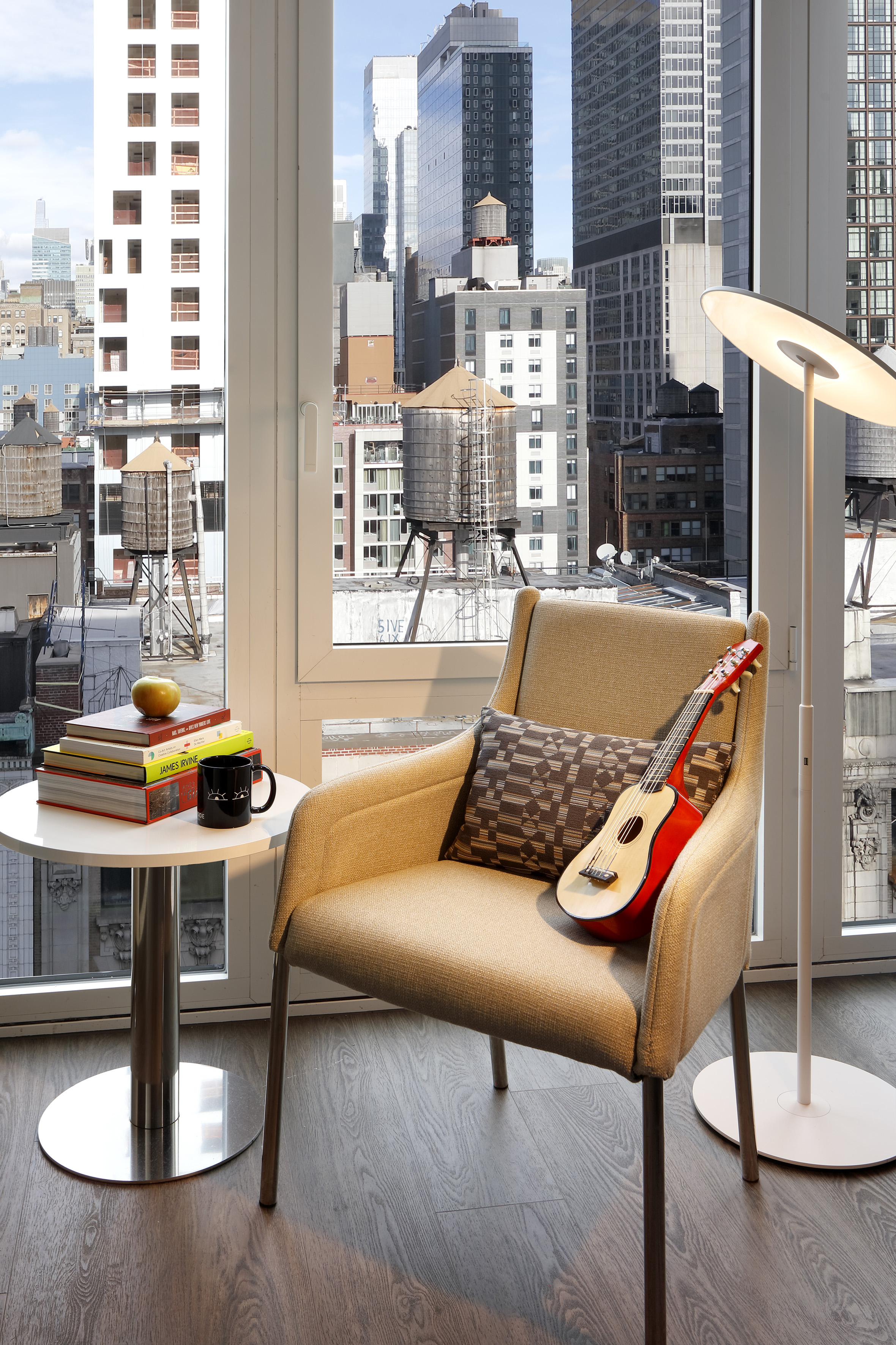 Modern chair with ukulele, books, and urban cityscape view from window.