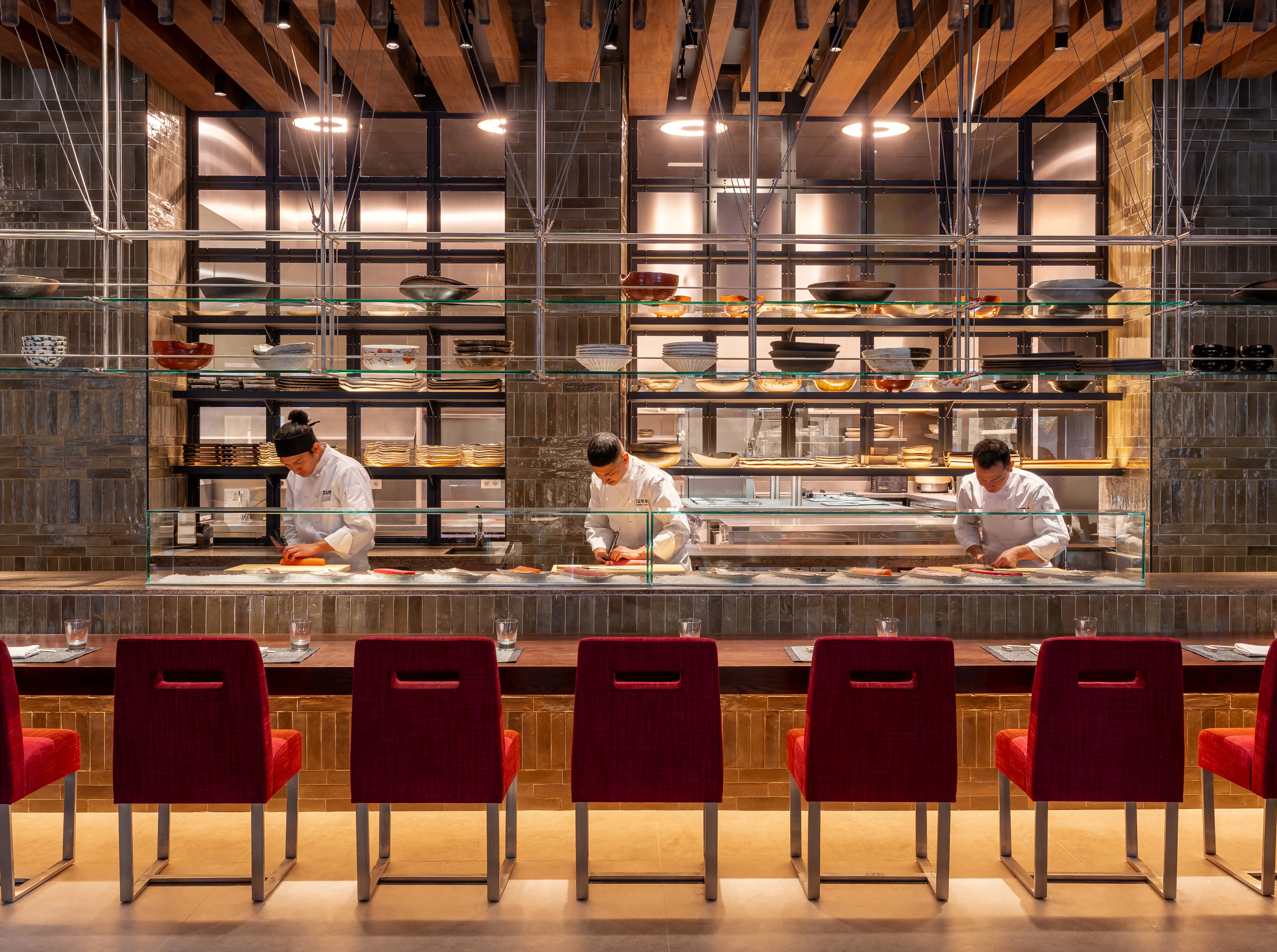 Chefs preparing sushi at a modern open kitchen sushi bar with red counter seating.