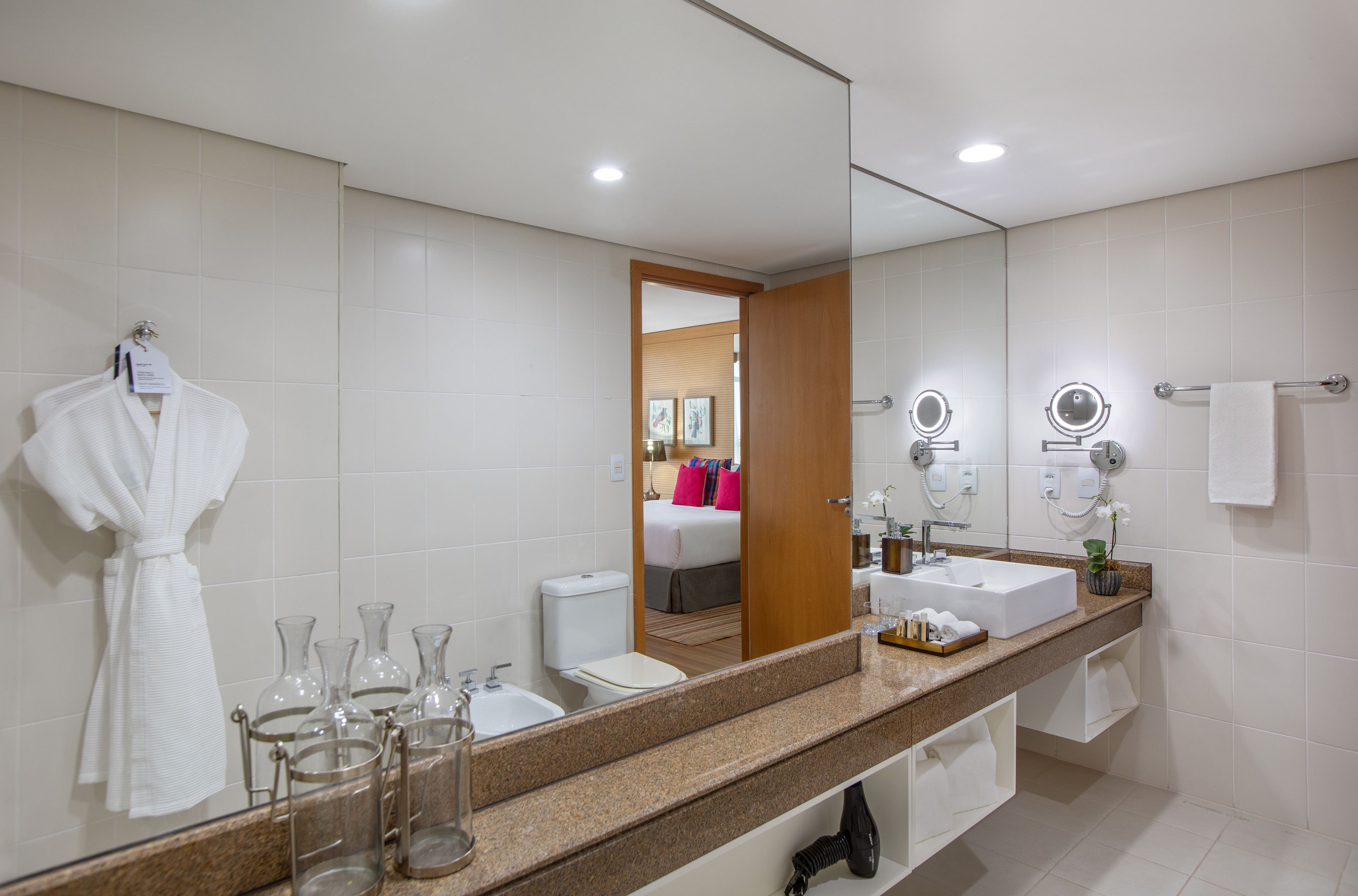 Modern hotel bathroom with vessel sink, granite counter, white robe, and bedroom view.