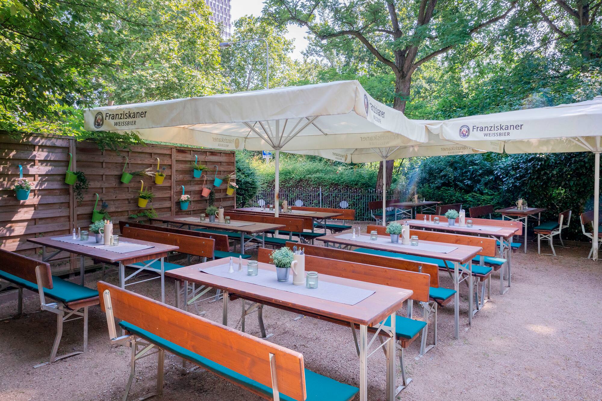 Jardín de cerveza con mesas y bancos, sombrillas Franziskaner y árboles.