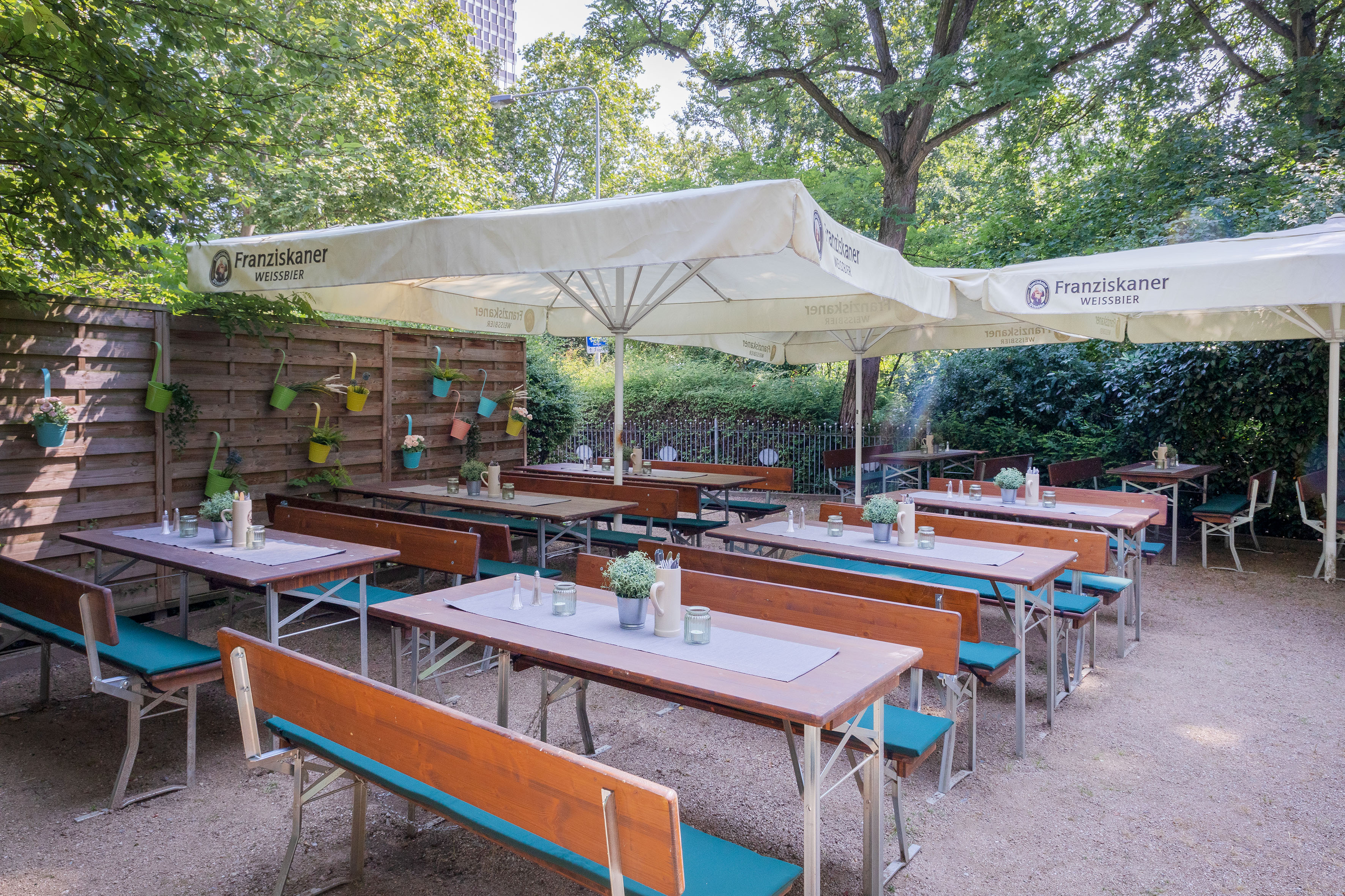 Jardín de cerveza con mesas y bancos, sombrillas Franziskaner y árboles.