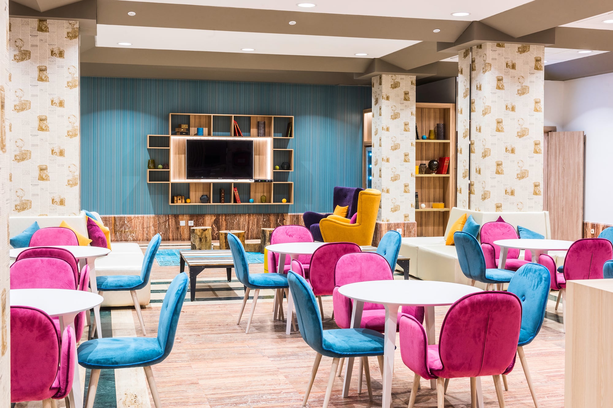 Modern vibrant lounge with pink and blue seating, white tables, and TV.