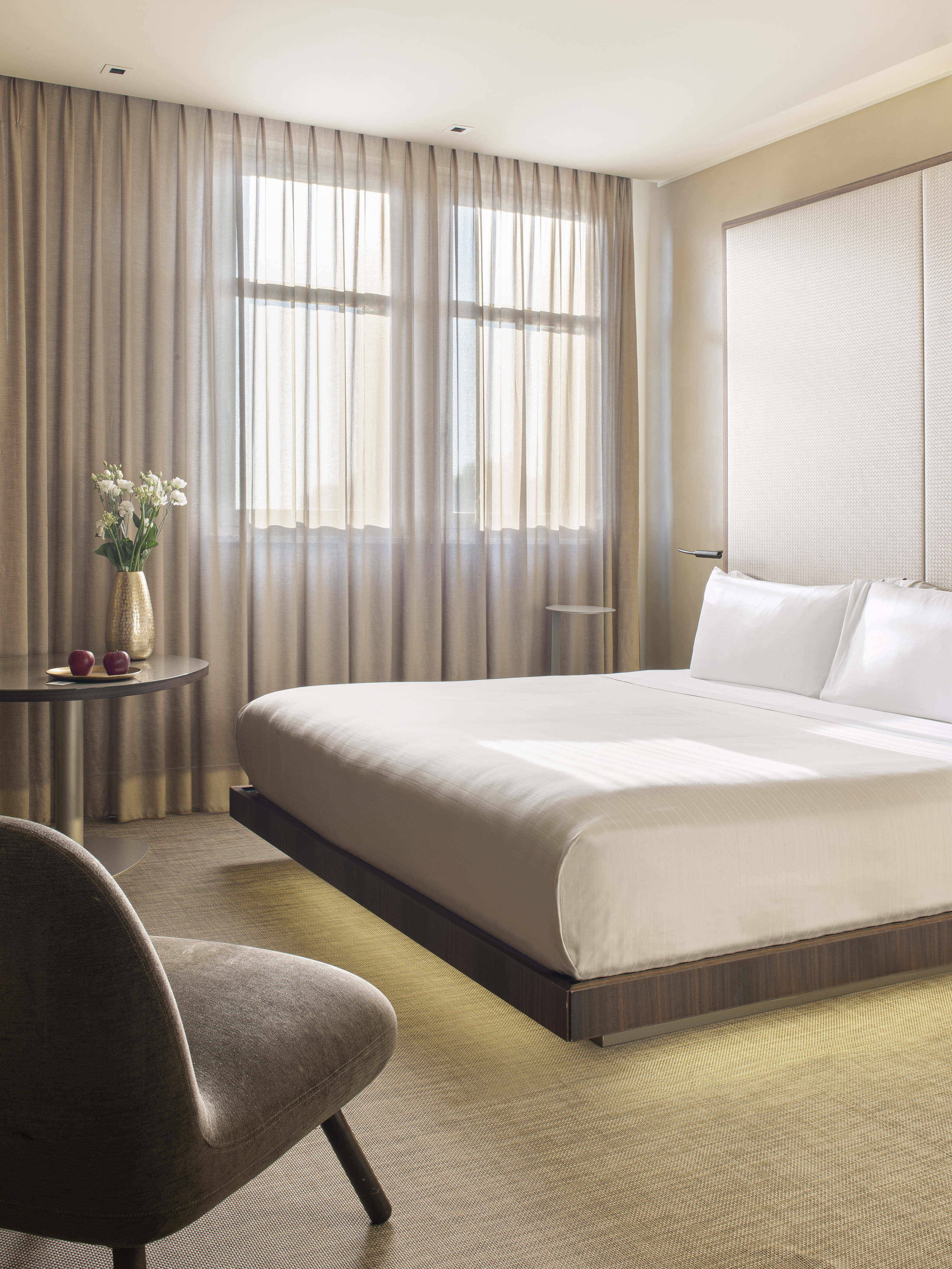 Modern hotel room with bed, sheer curtains, side table, and flowers.