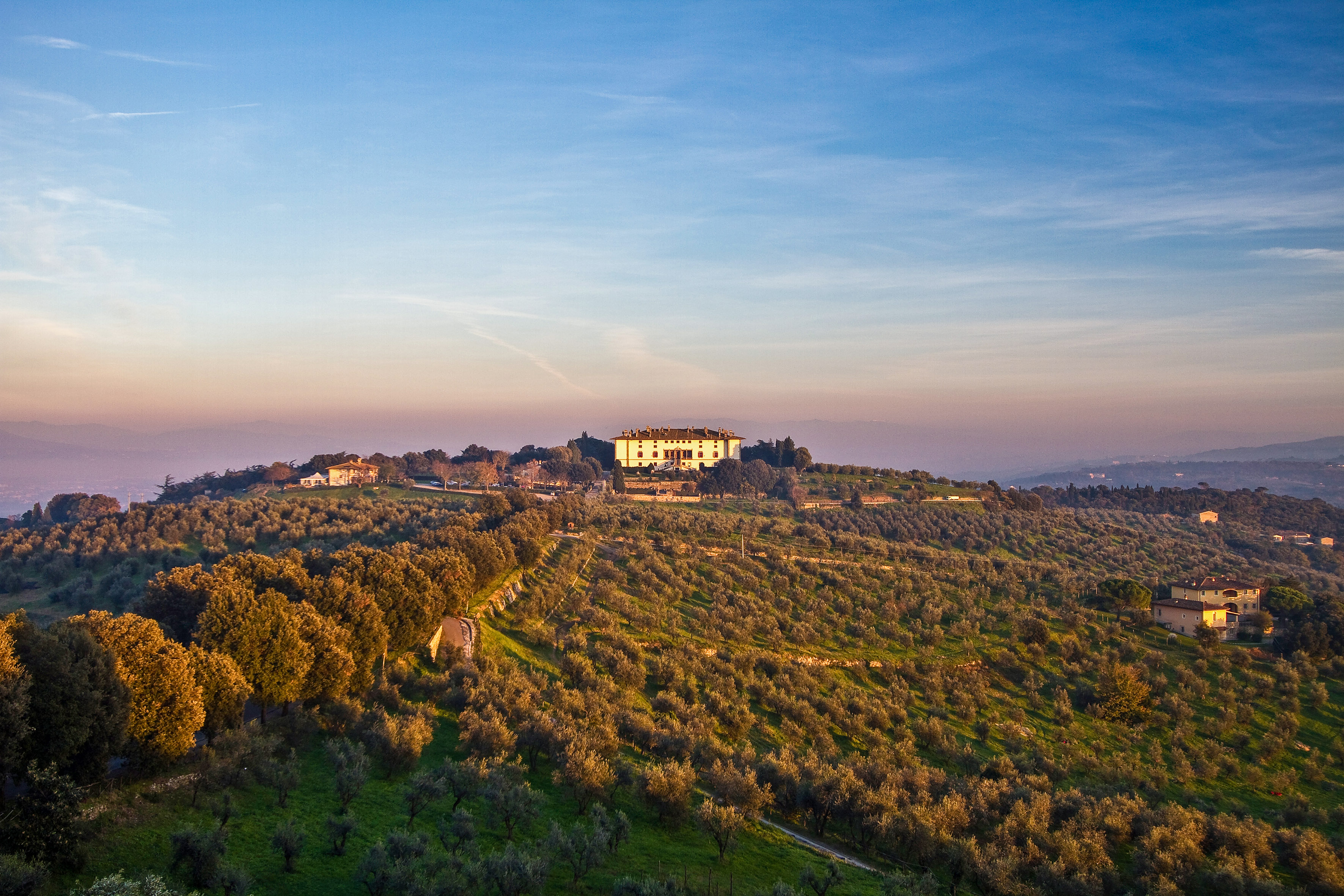 White villa on a Tuscan hilltop surrounded by sunlit olive groves at sunset.