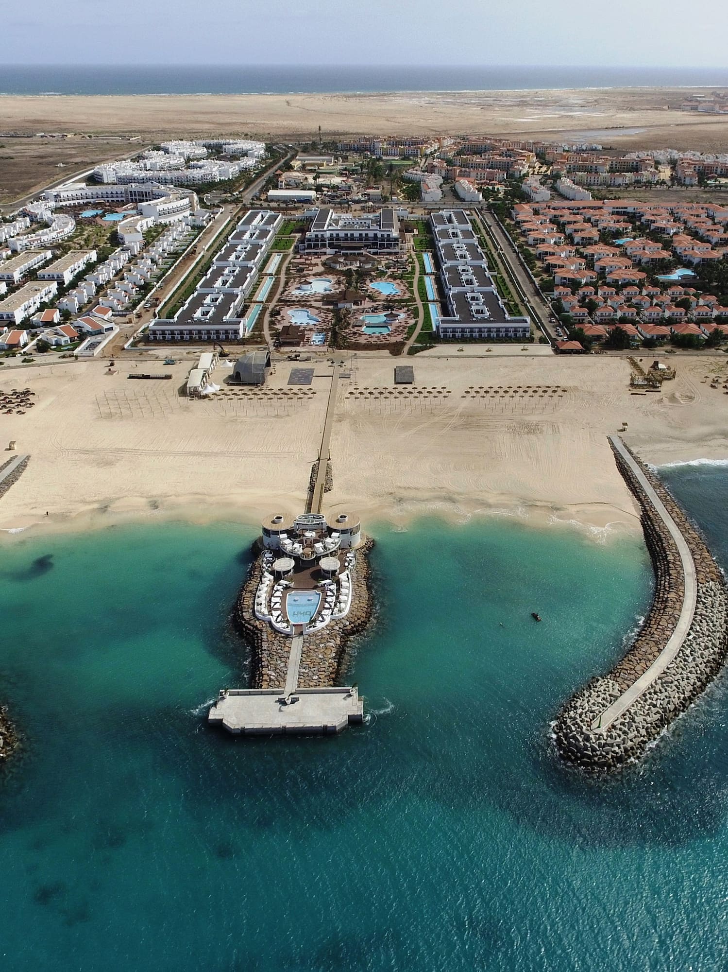 Aerial: beachfront resort, sandy beach, turquoise ocean, crescent breakwaters.