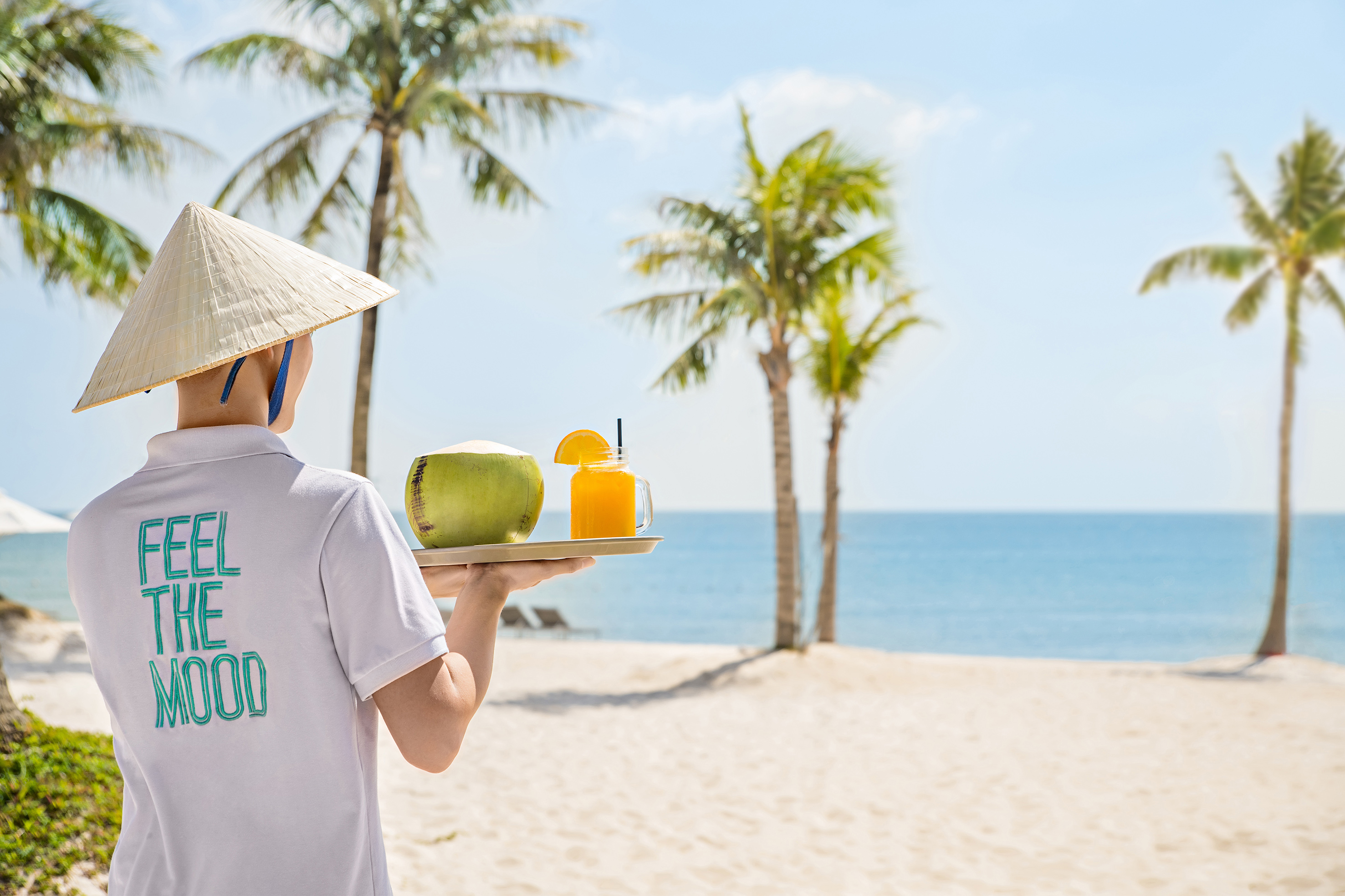 Server in conical hat brings tropical drinks on a sunny beach.