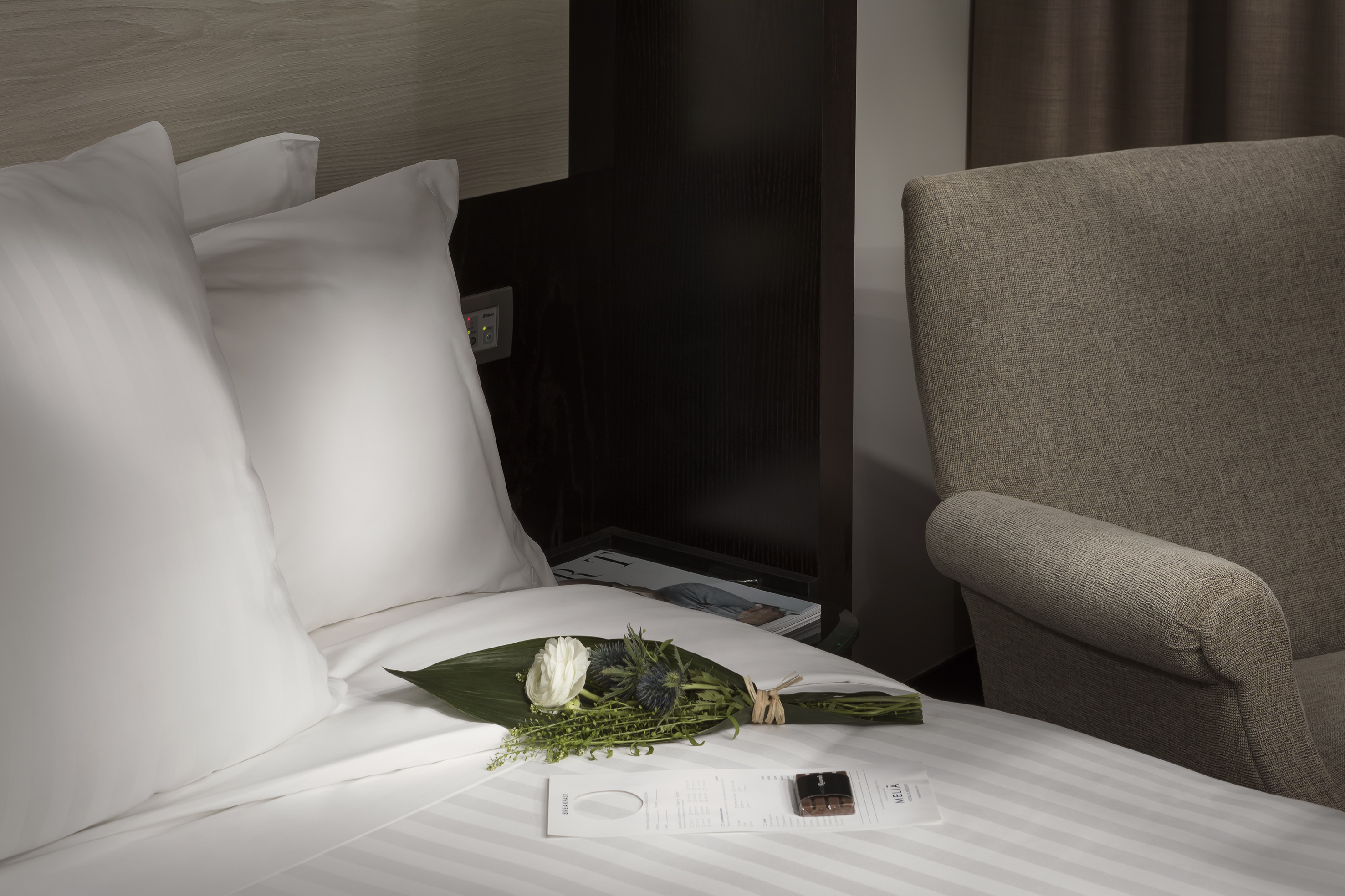 Elegant hotel bed with welcome flowers, Meliá chocolate, and breakfast menu.