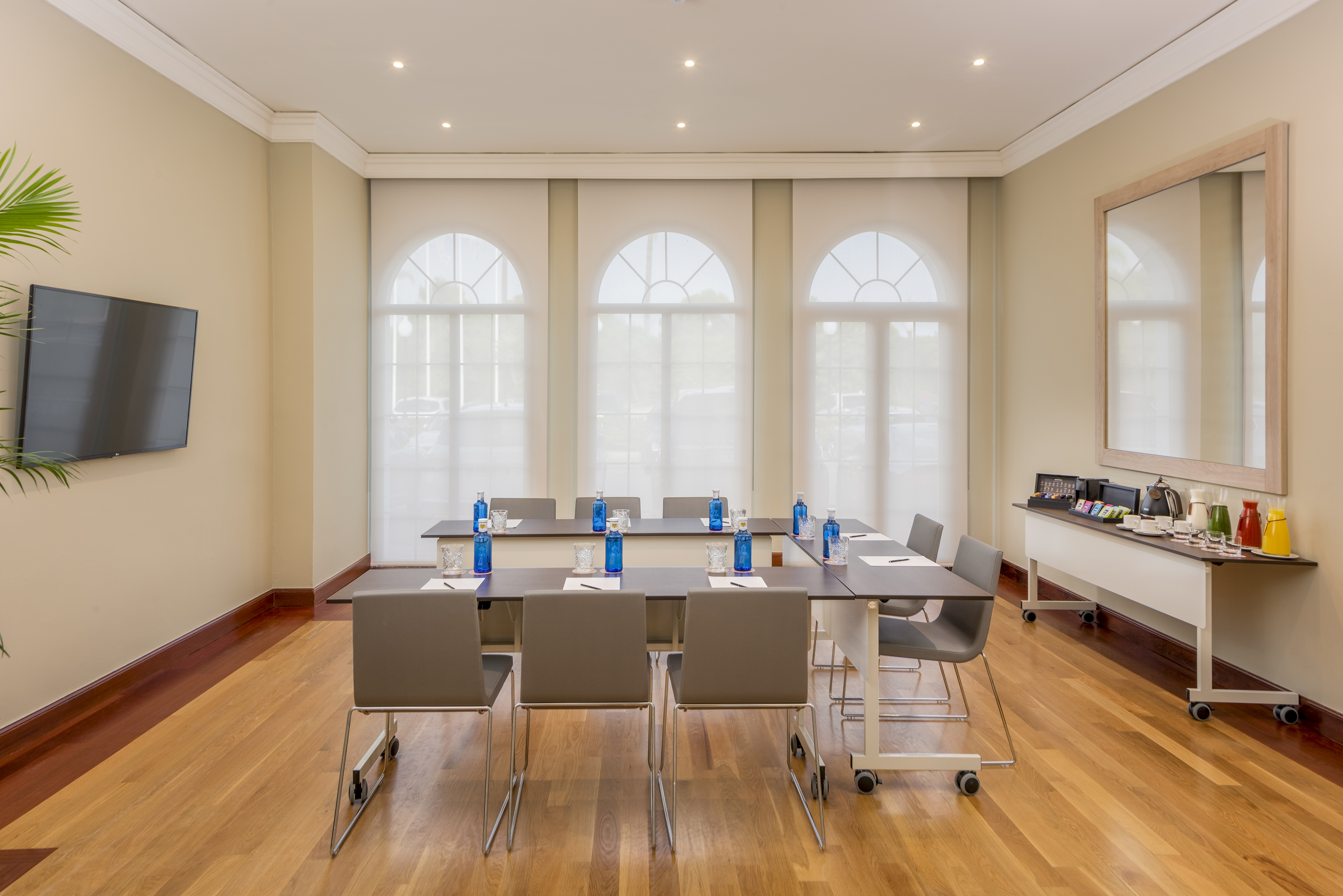 Modern meeting room with U-table setup, TV, hardwood floor, and arched windows.