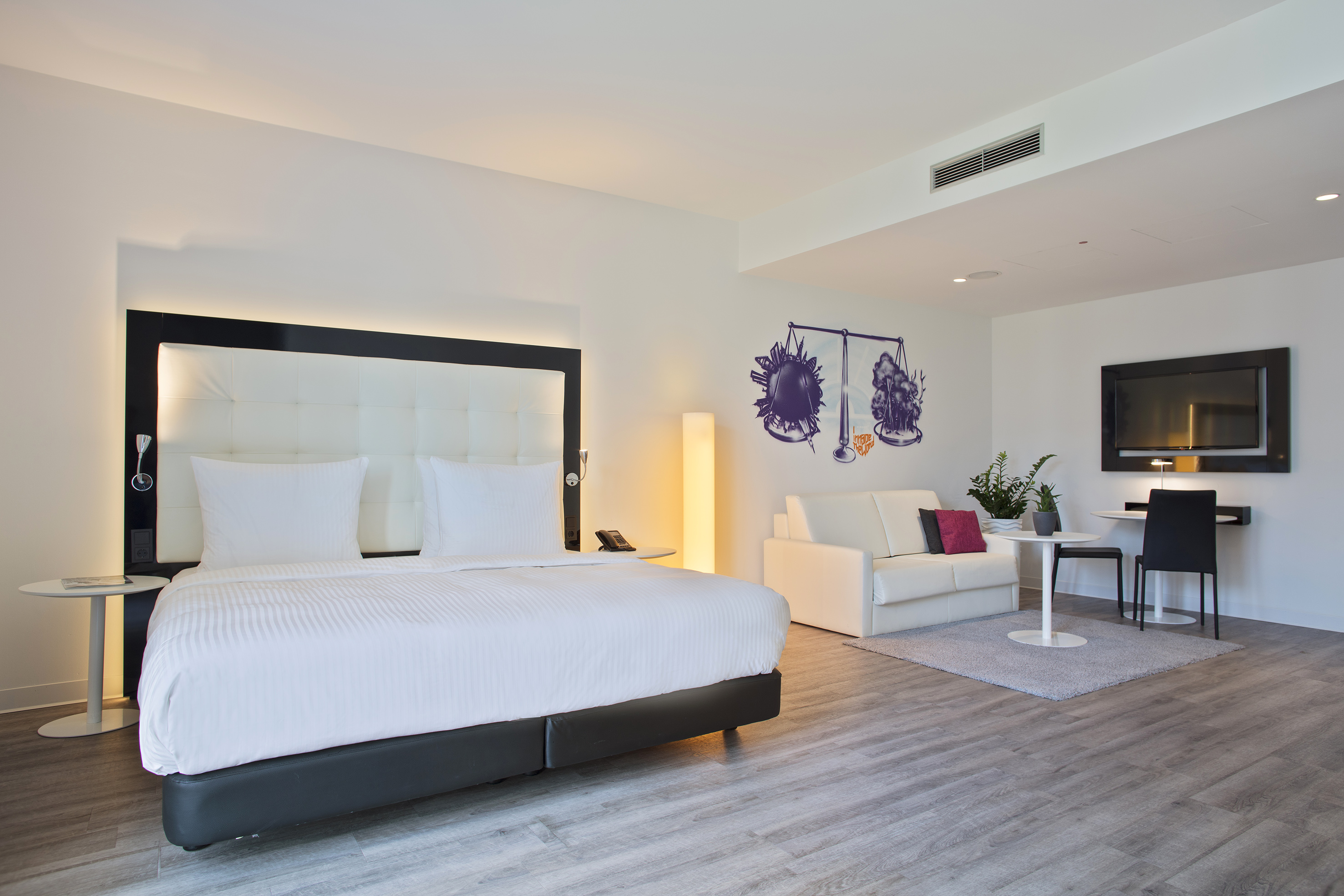 Modern hotel room with large bed, white sofa, TV, and desk area.