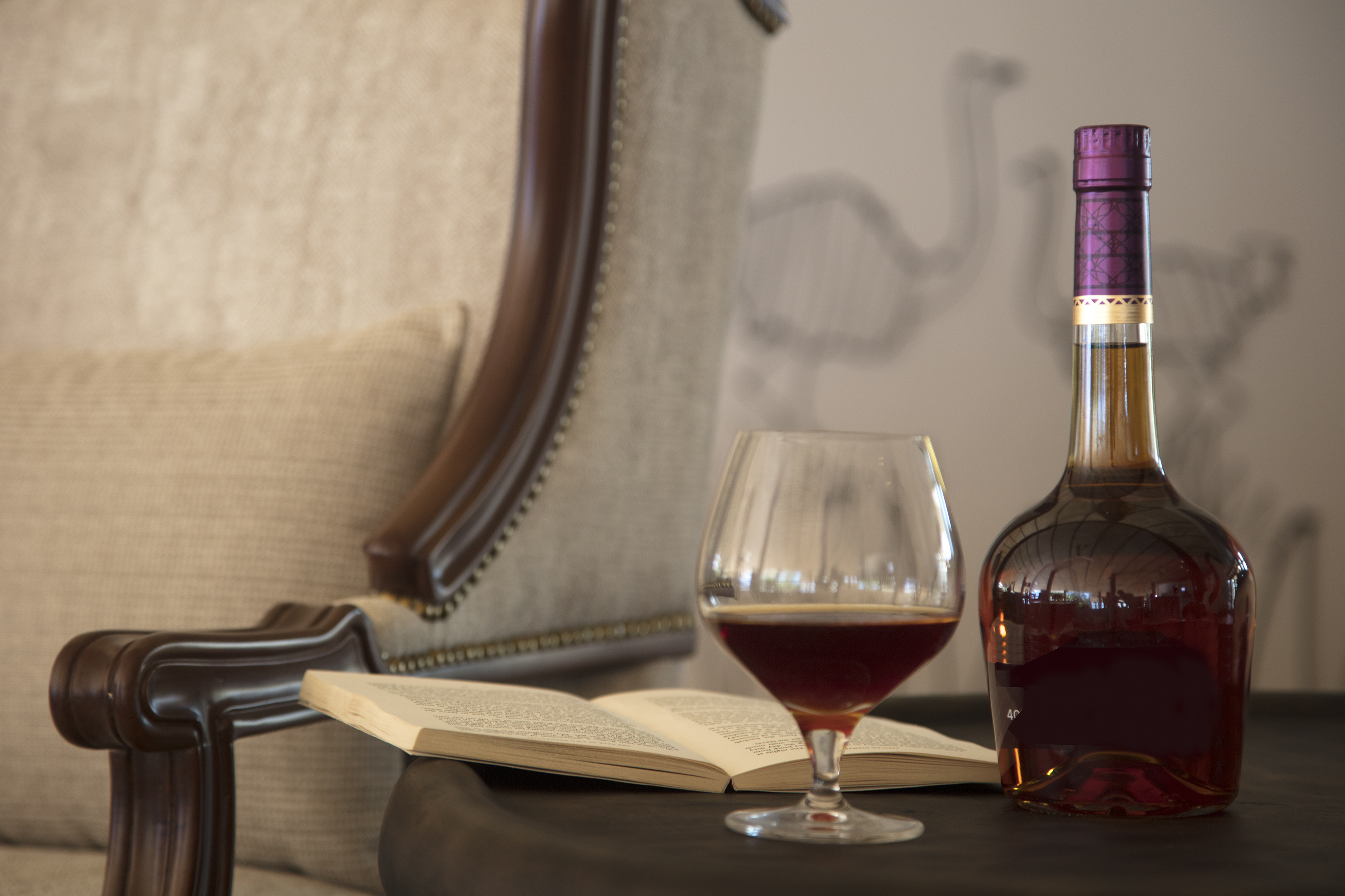 Brandy bottle, snifter glass, and open book on table by armchair.