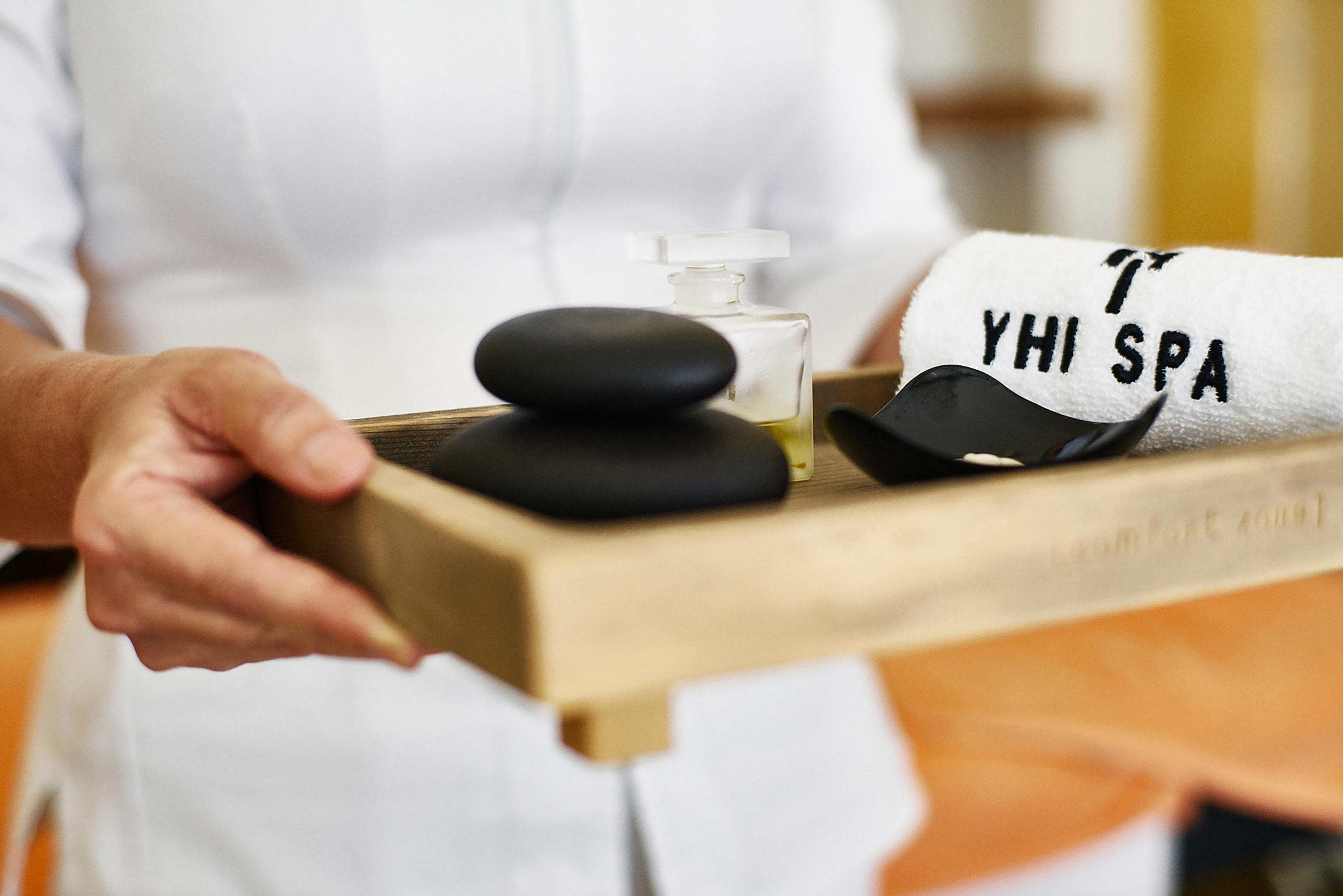 Hands holding spa tray with hot stones, essential oil, and YHI SPA towel.