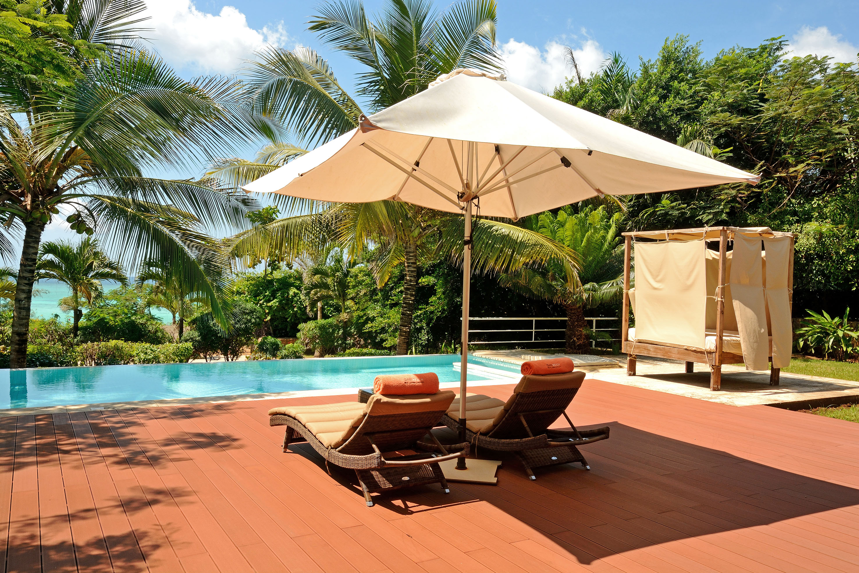 Tropical resort pool deck with lounge chairs, umbrella, palm trees, ocean view.