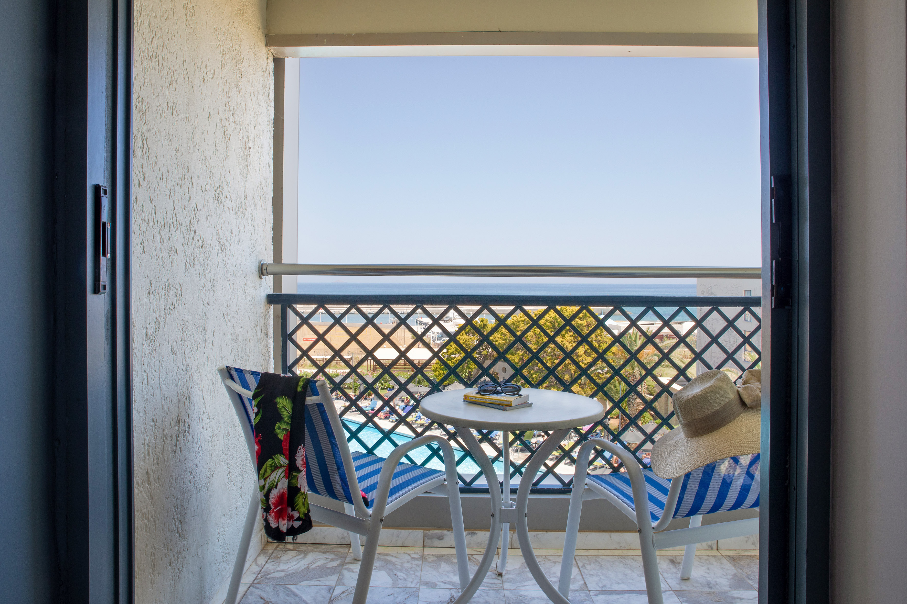 Sunny hotel balcony with ocean view, resort pool, striped chairs, table.