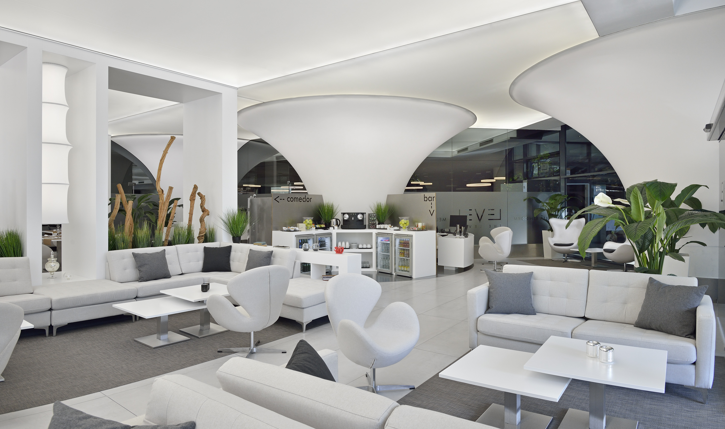 Modern hotel lounge with white sofas, snack bar, plants, and curved ceilings.