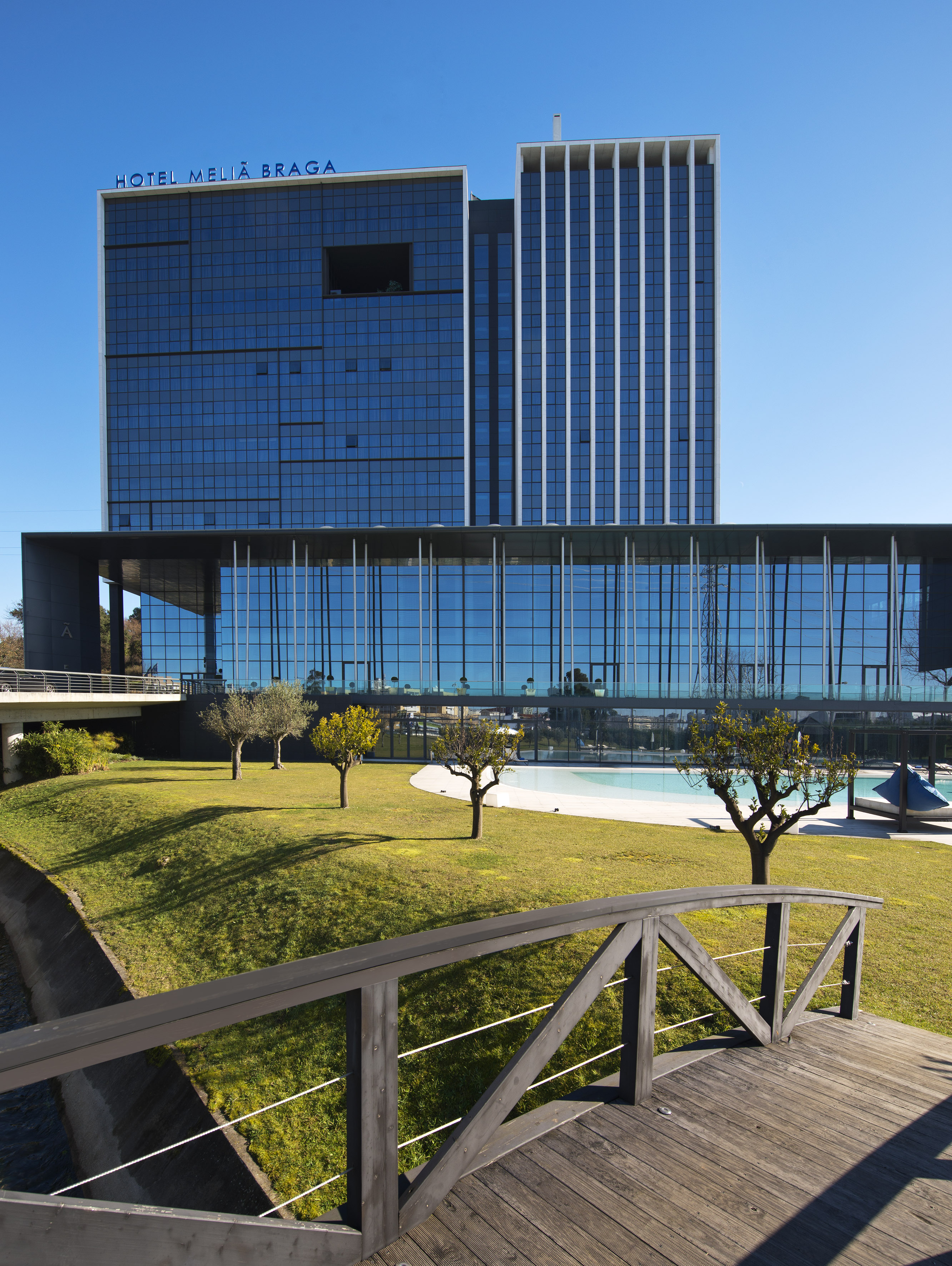 Meliã Braga hotel, modern glass building, garden, pool, and wooden bridge.